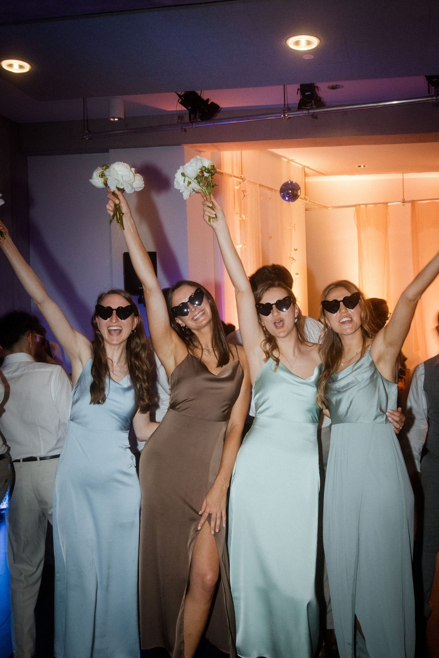Bridesmaids in pastel dresses hold flowers overhead while posing with sunglasses indoors.