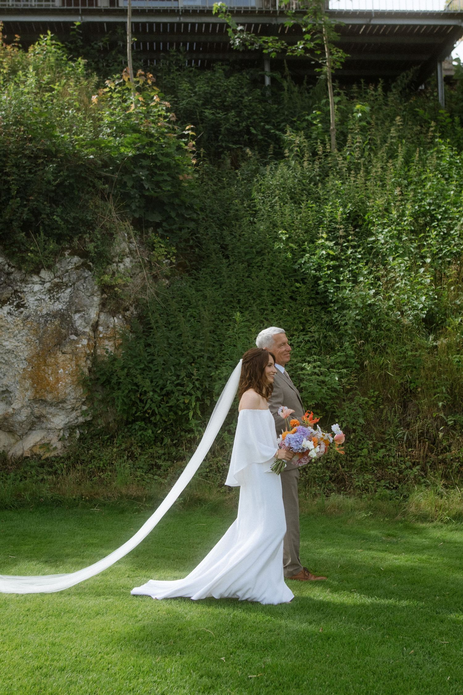 Wedding couple in formal attire posing in a lush garden setting with flowing veil.