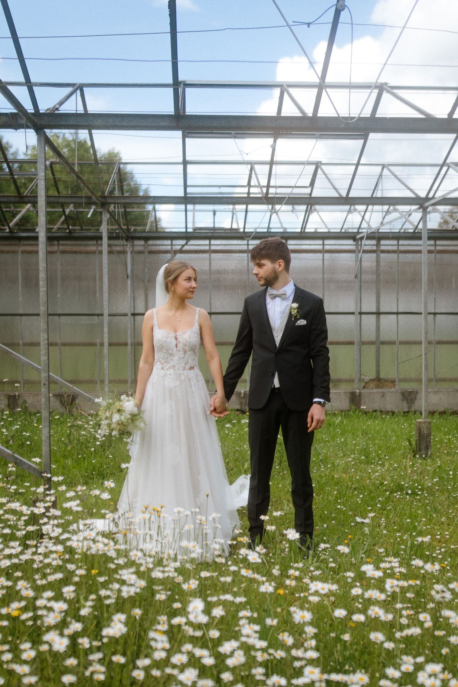 Wedding couple stands together in a greenhouse surrounded by white wildflowers.