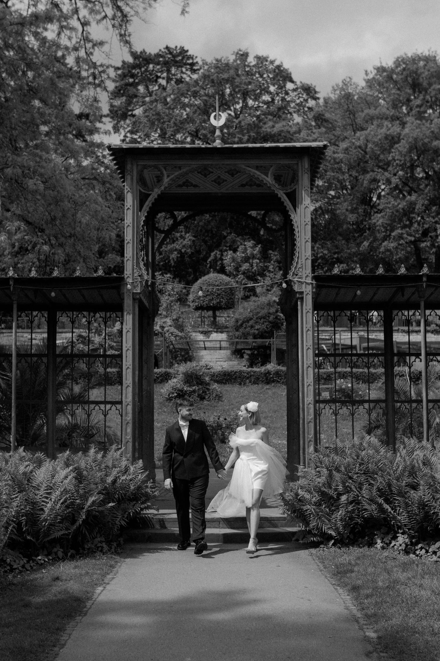 Black and white photo of newlyweds walking through decorative garden entrance.