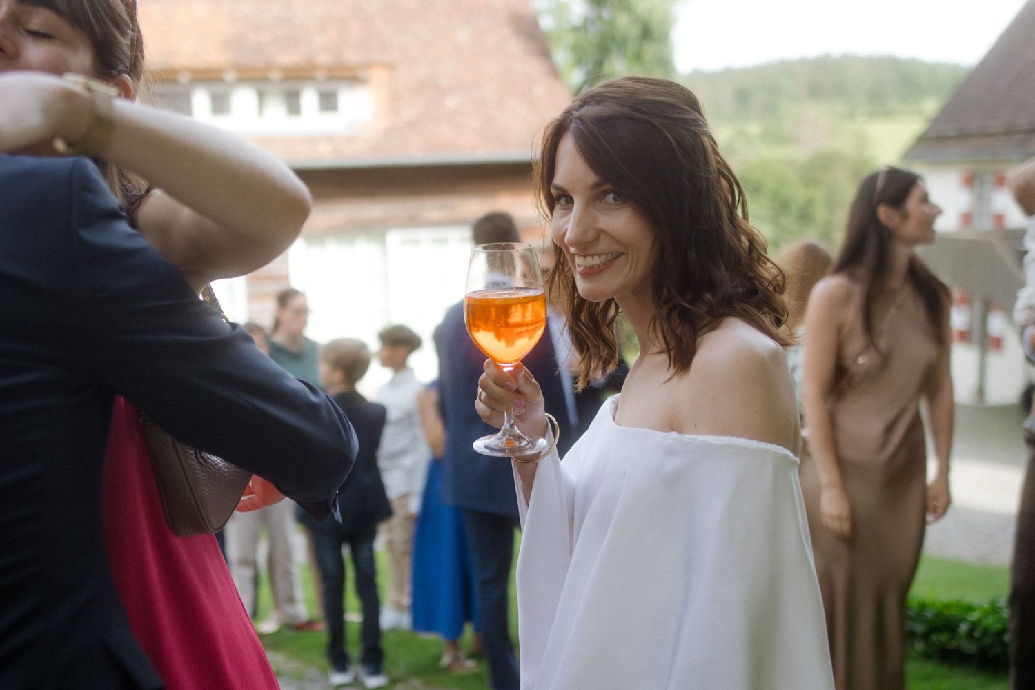 A wedding celebration guest in white dress holds an aperol spritz cocktail outdoors.