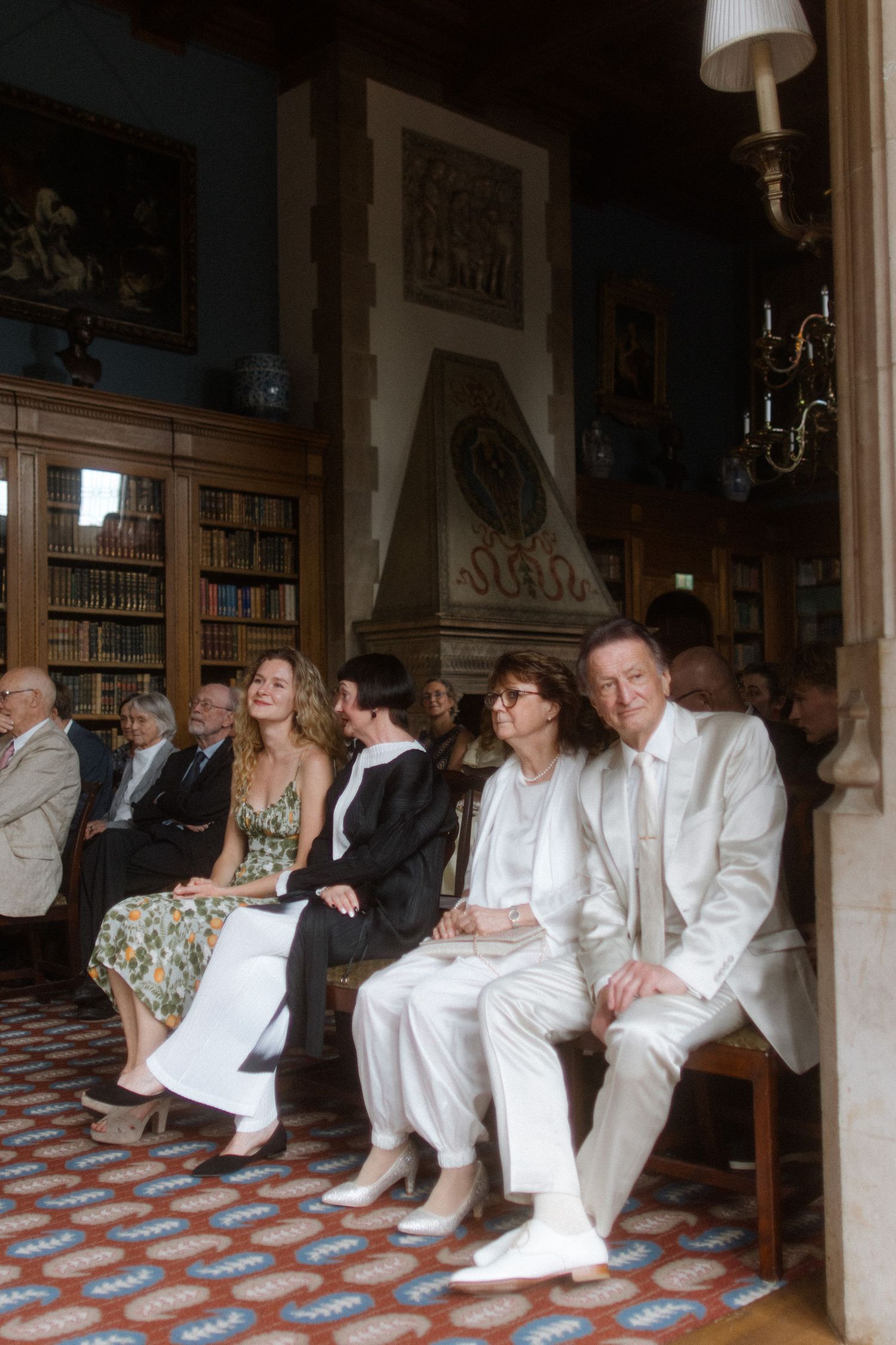 Wedding guests in white attire seated in a grand library with checkered floors.