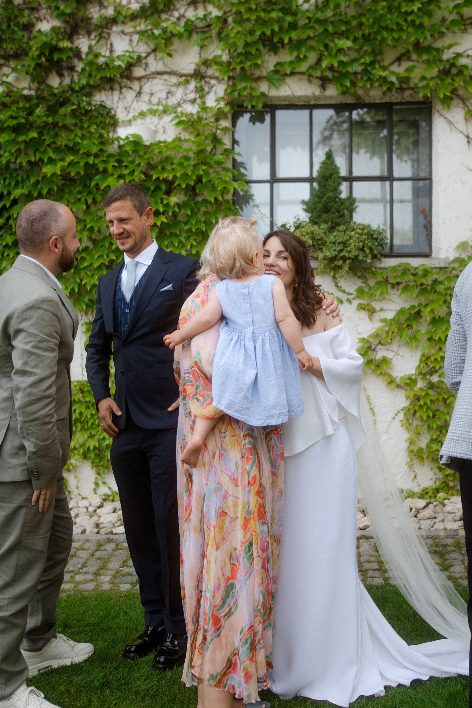 Wedding party gathers for candid moments in front of ivy-covered wall during outdoor ceremony.