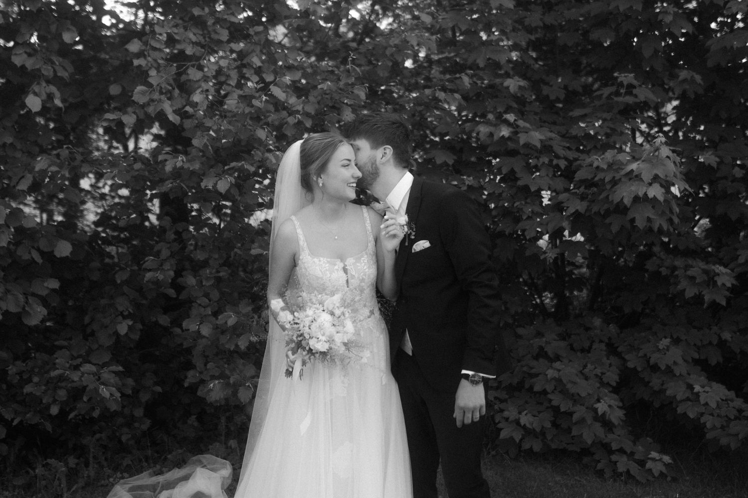 A romantic black and white wedding portrait captures a tender moment between newlyweds against a leafy garden backdrop.