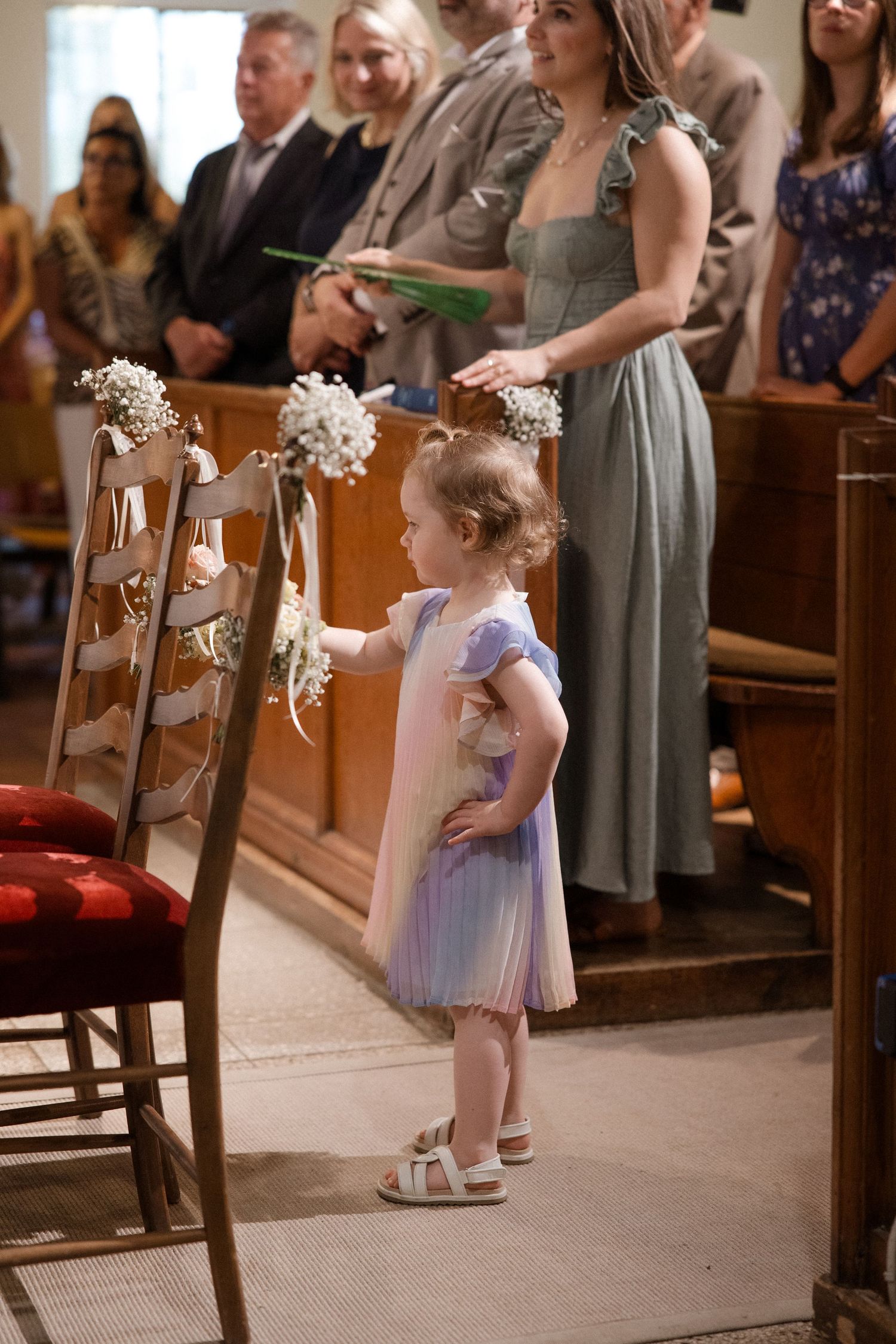 Small flower girl in lavender dress stands beside pew decorated with white flowers.