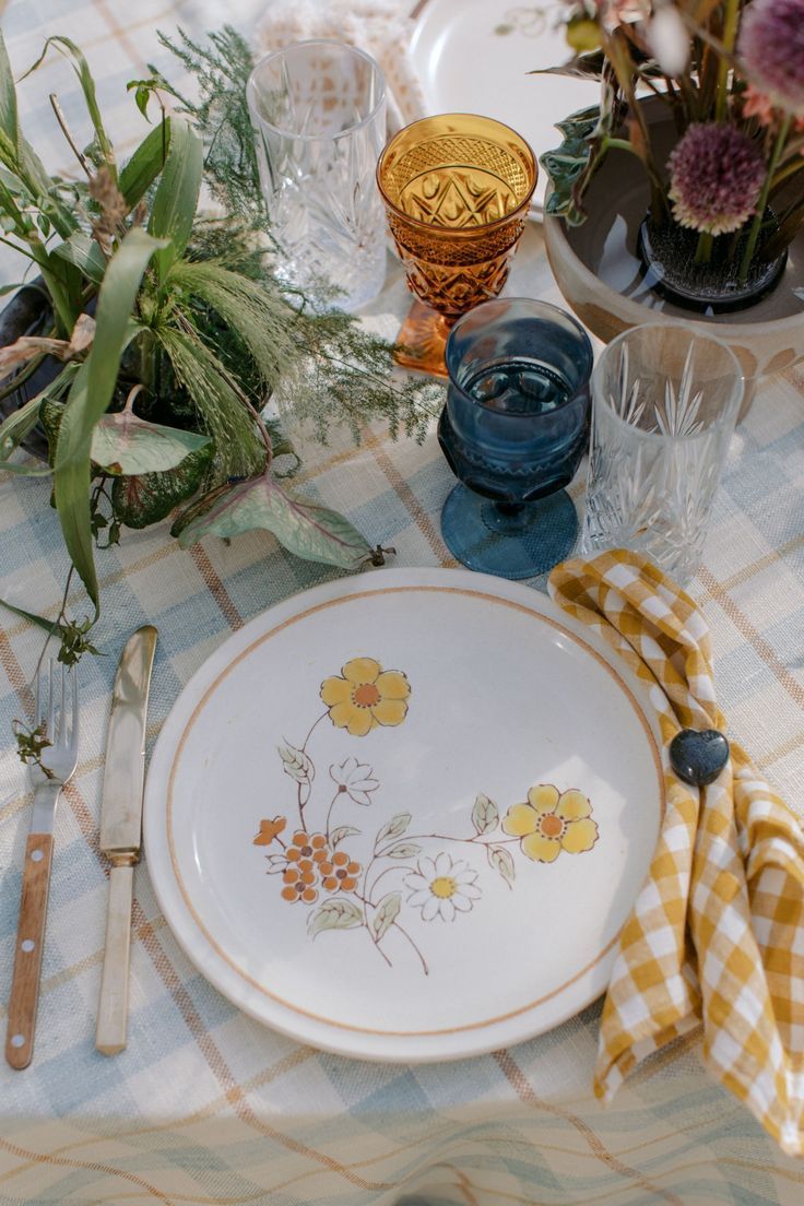Place setting with floral plate, colored glassware, and yellow gingham napkin on checkered tablecloth.
