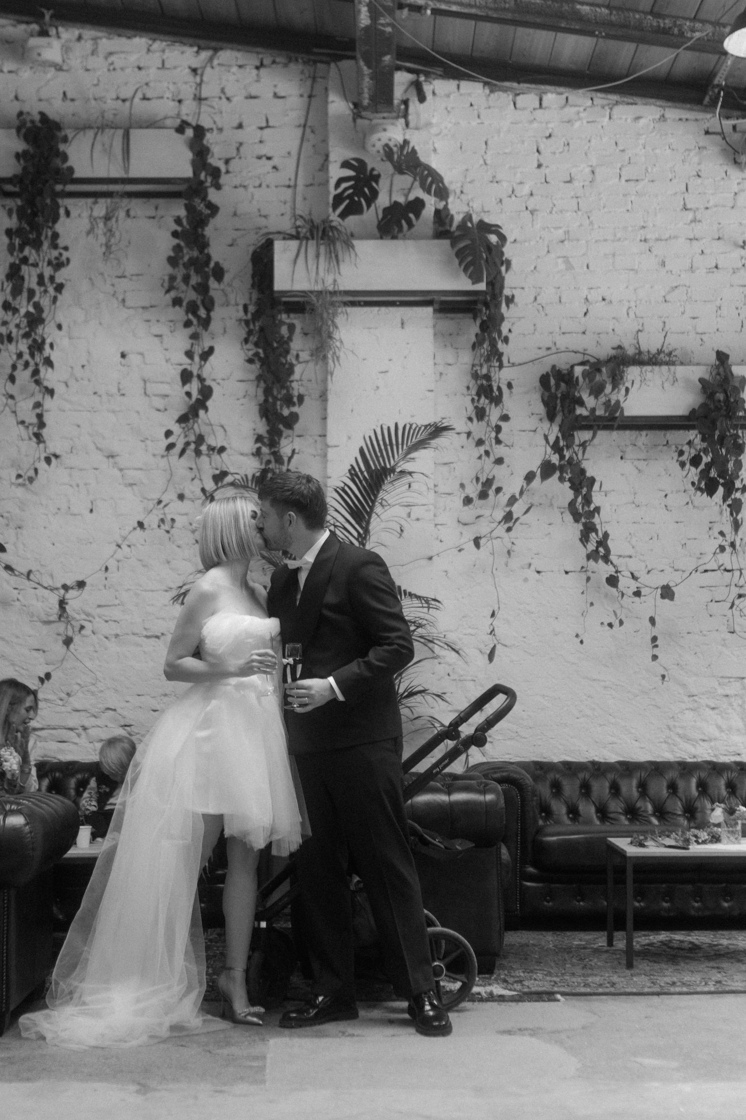 A couple shares an intimate moment against a white brick wall decorated with cascading vines.