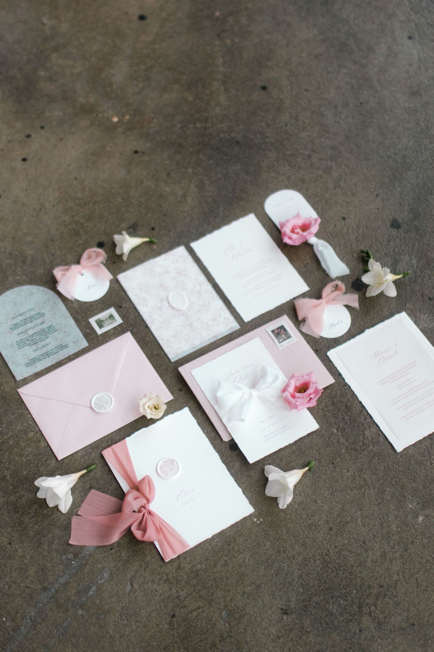 Wedding invitation collection featuring pink roses and white paper elements on concrete surface.