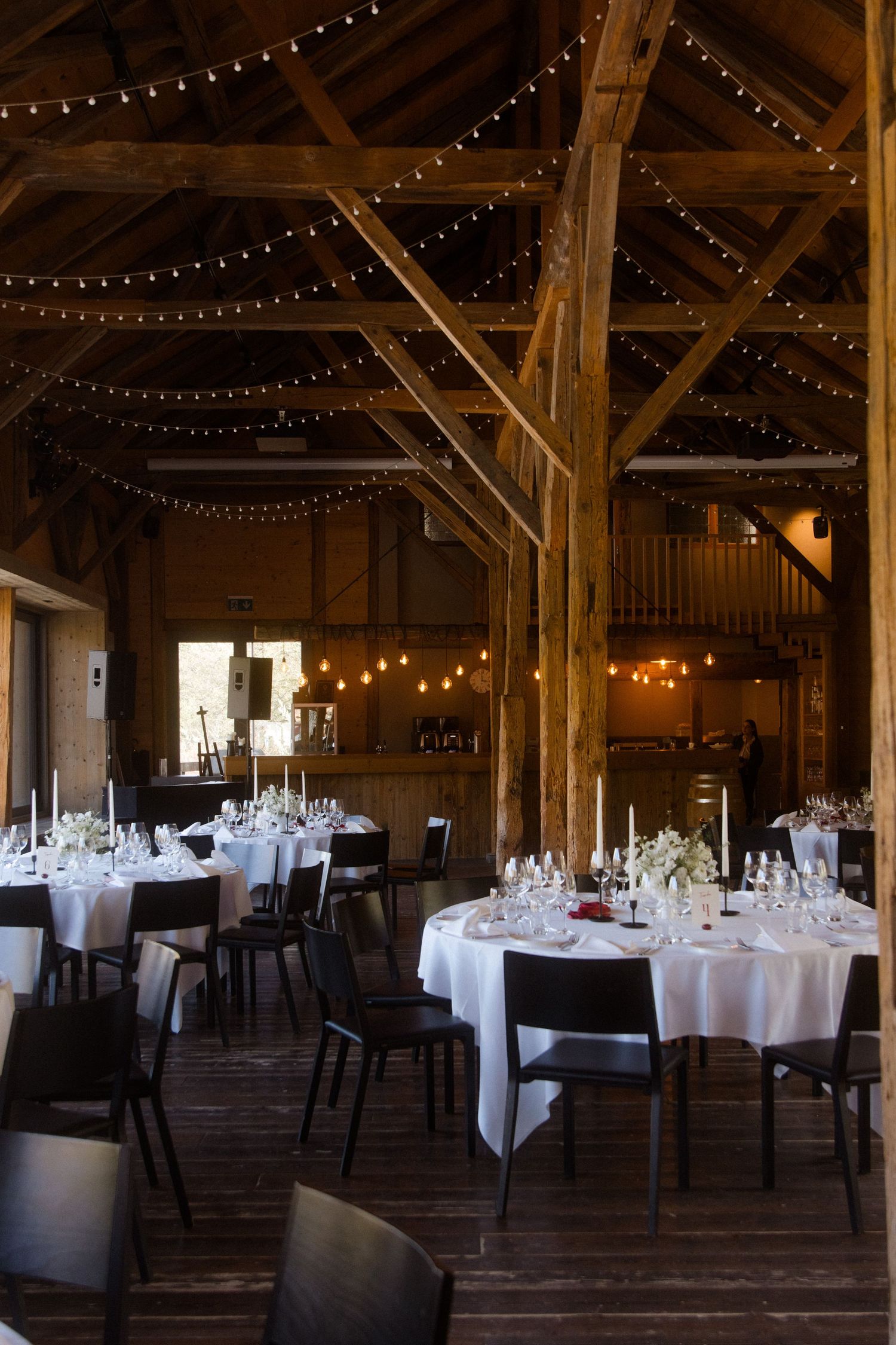 Rustic barn wedding reception with string lights, wooden beams, round tables with white linens and candles.