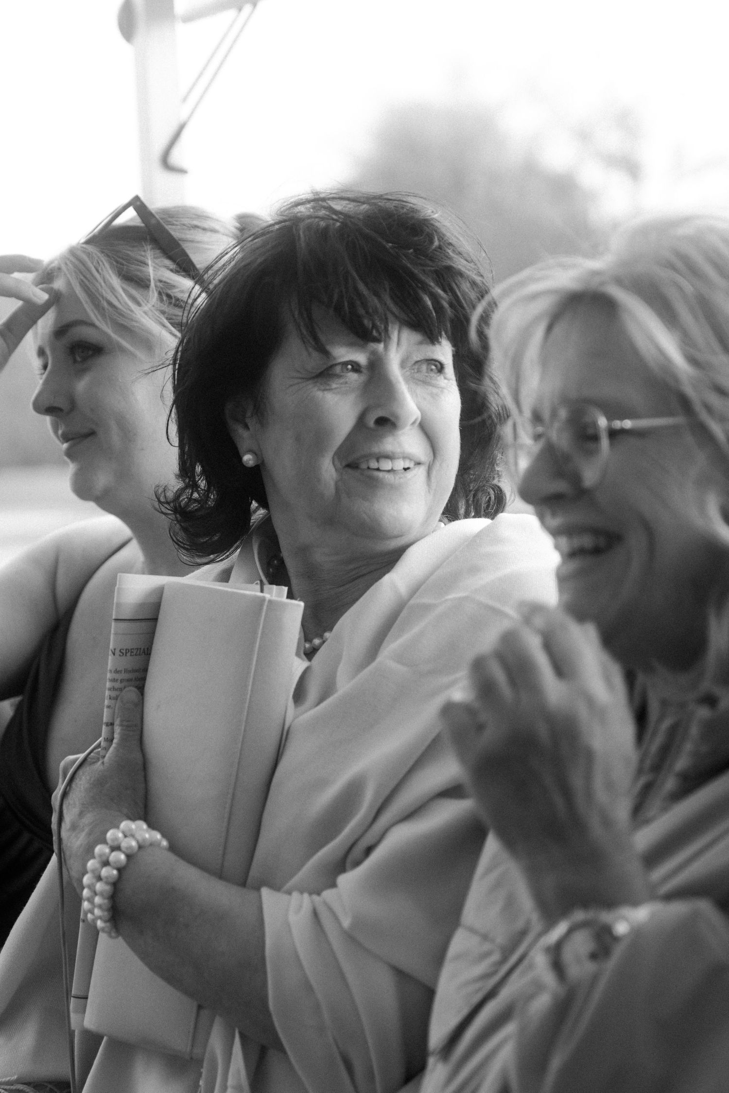 Three women smile and laugh together at an outdoor wedding celebration in black and white photo.