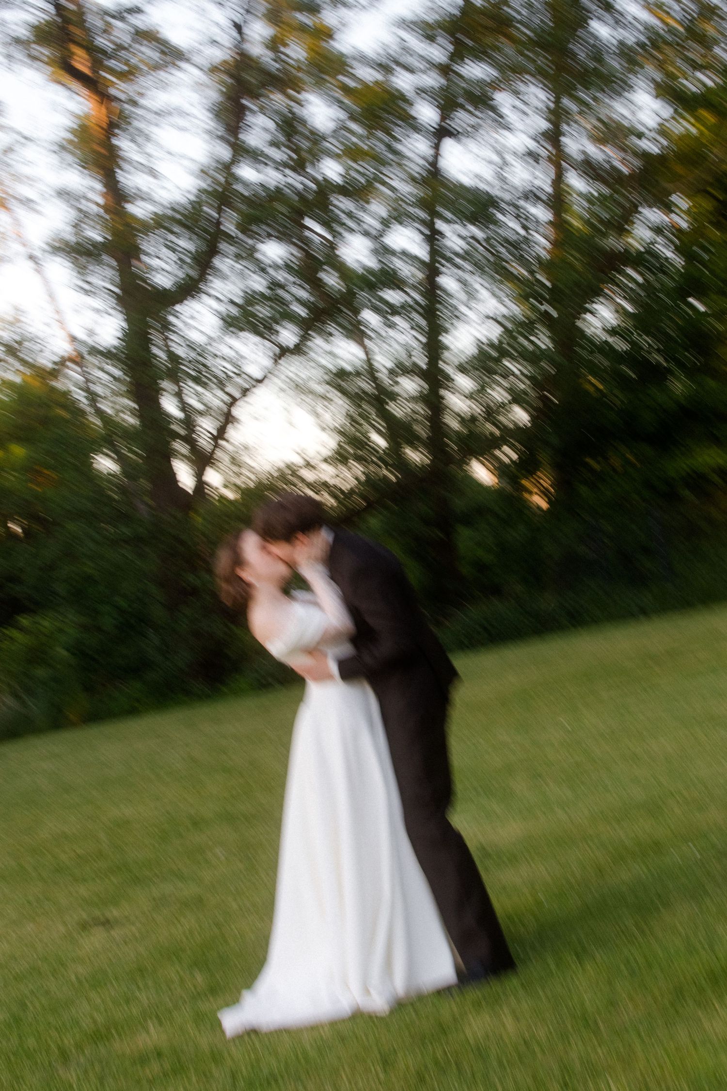 Blurred motion photo of bride and groom sharing a romantic kiss on green grass with trees in background at sunset.