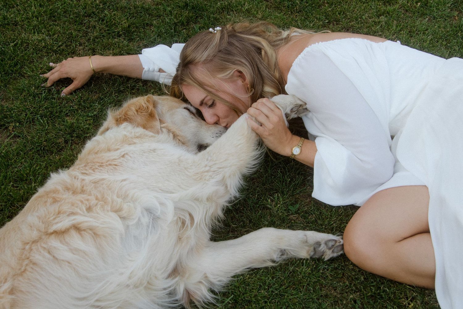 A person in white lying in grass next to a fluffy white dog.