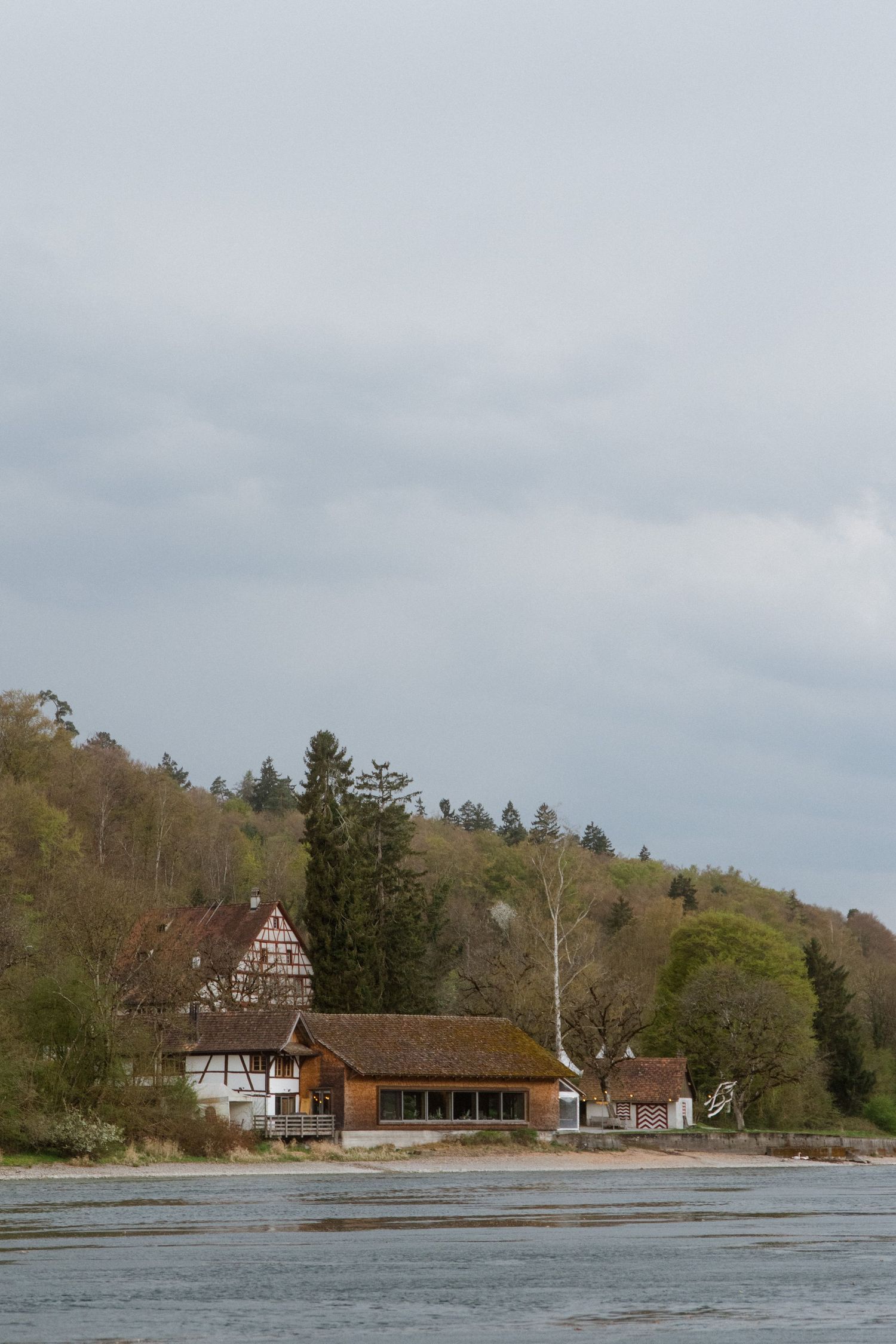 Rustic half-timbered house and wooden building beside a river with forested hills under cloudy sky.