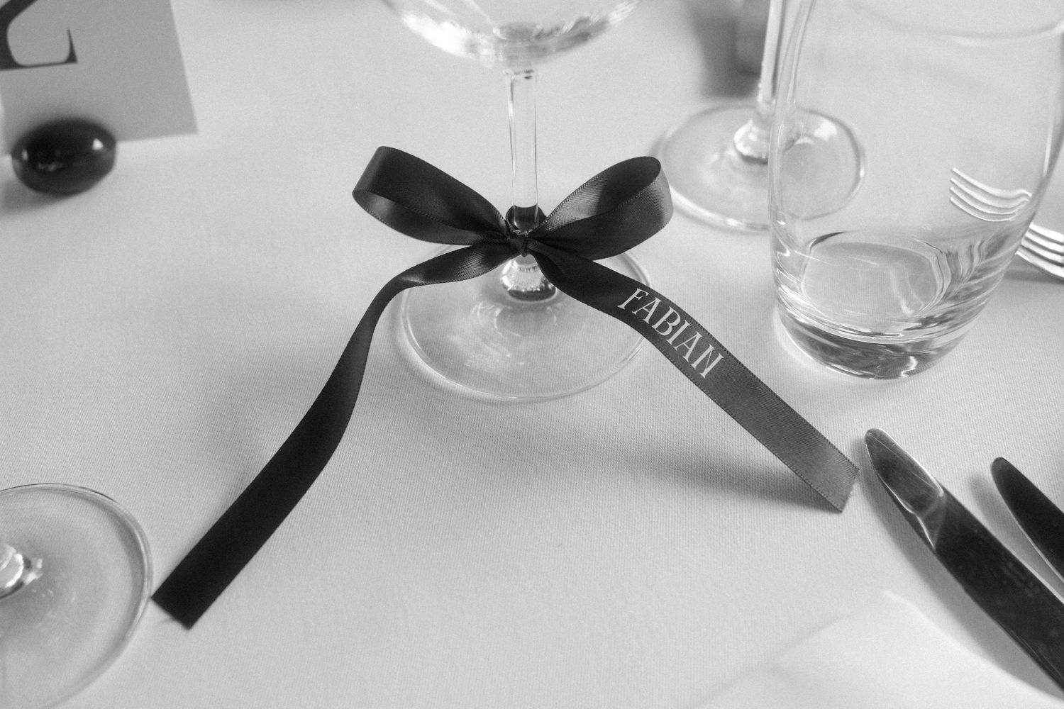 Black and white photo of an elegant place card tied with a black ribbon bow on a wine glass stem at a formal event.