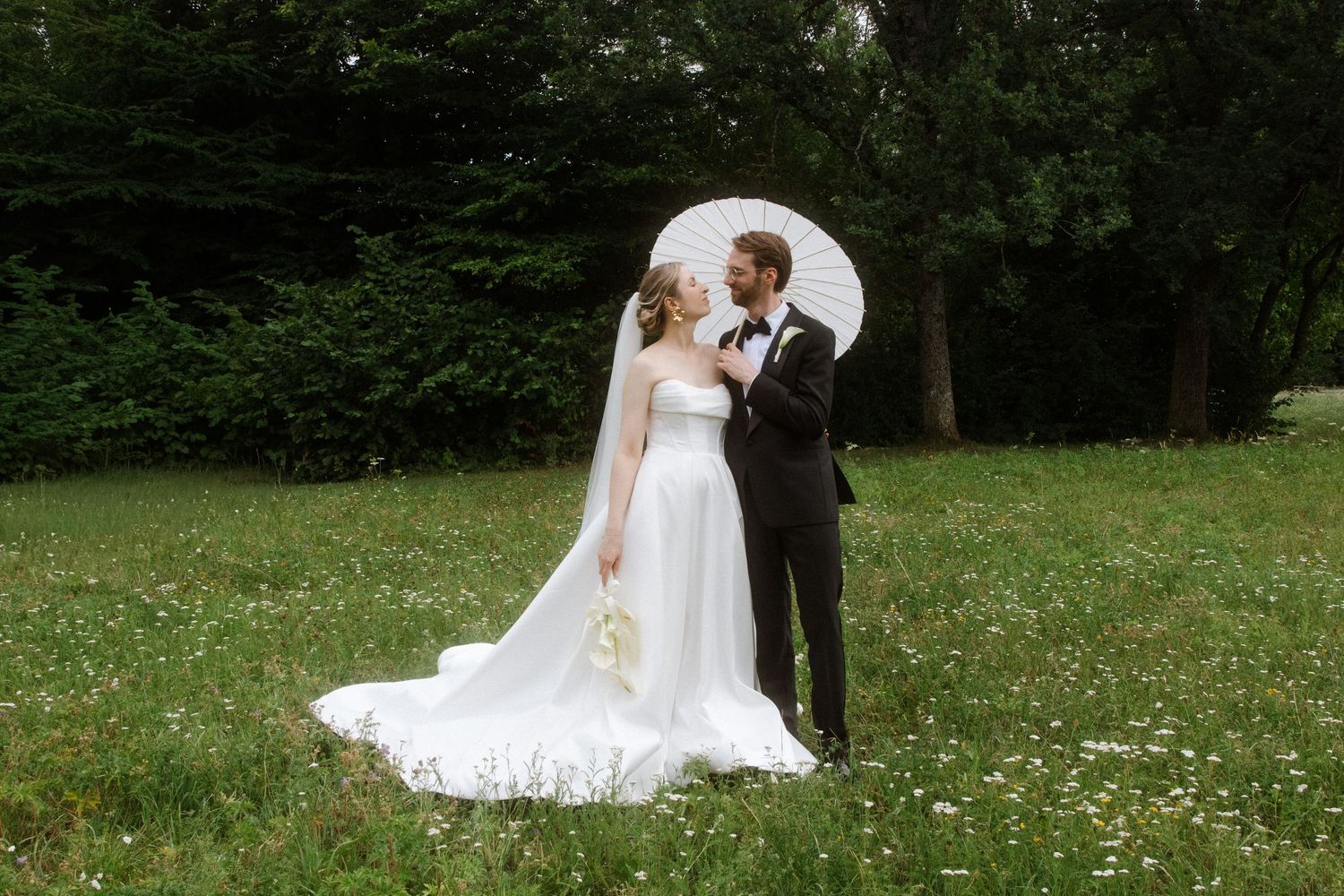 A couple takes wedding photos in a scenic meadow with trees in the background.