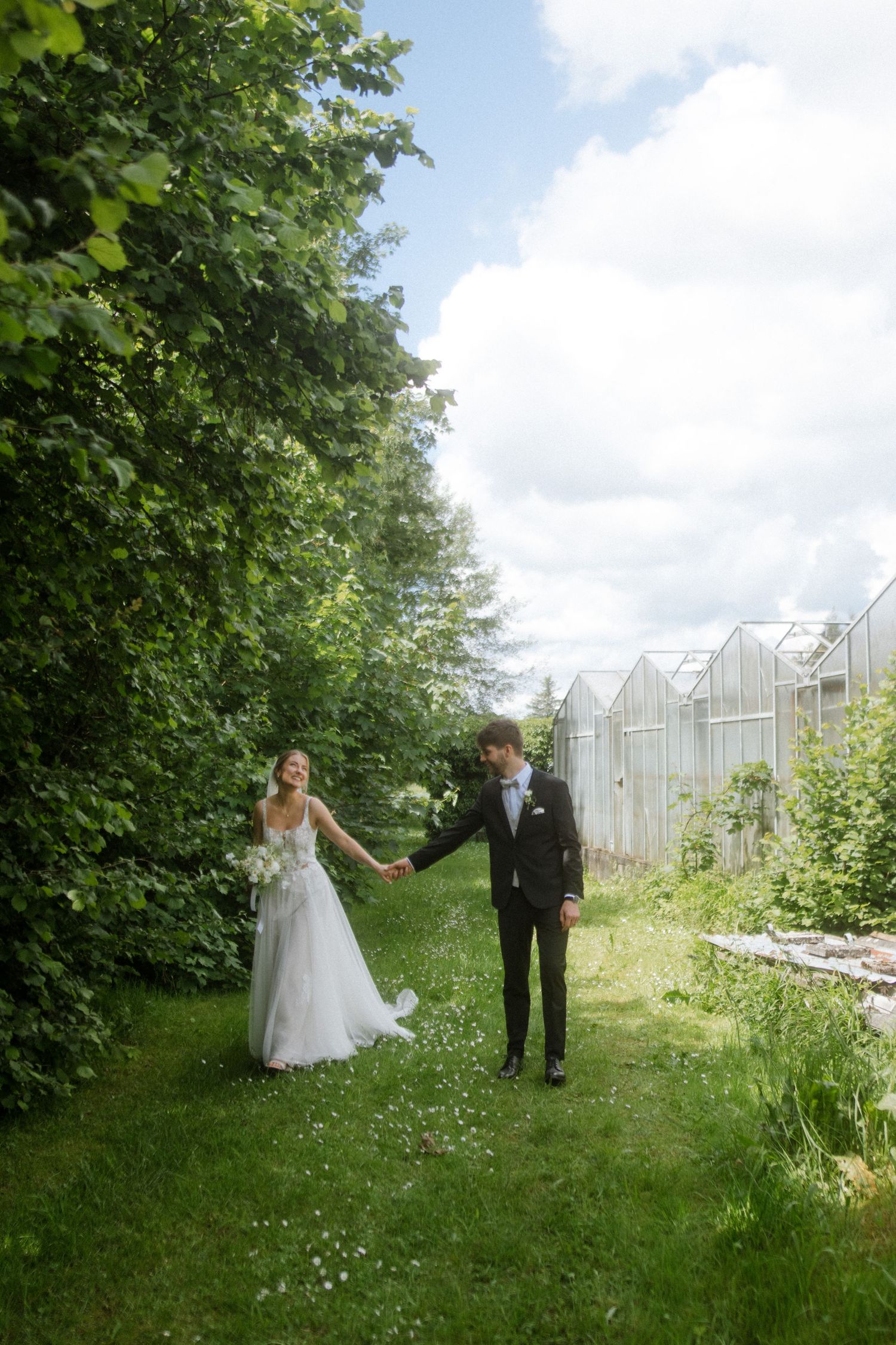 Newlyweds hold hands while walking along a grassy path beside greenhouse structures on their wedding day.