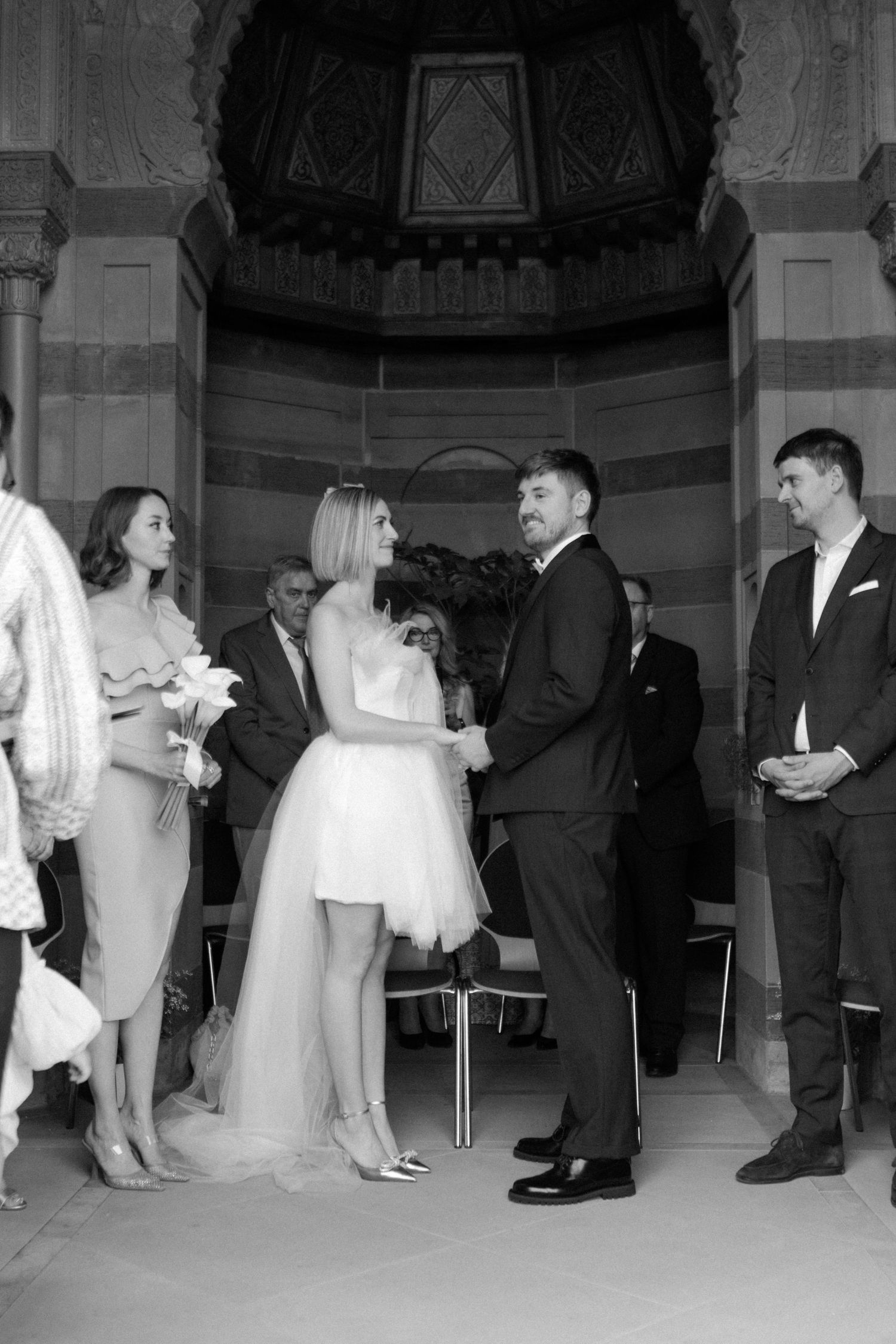 Intimate wedding ceremony in ornate alcove with wedding party gathered around couple.
