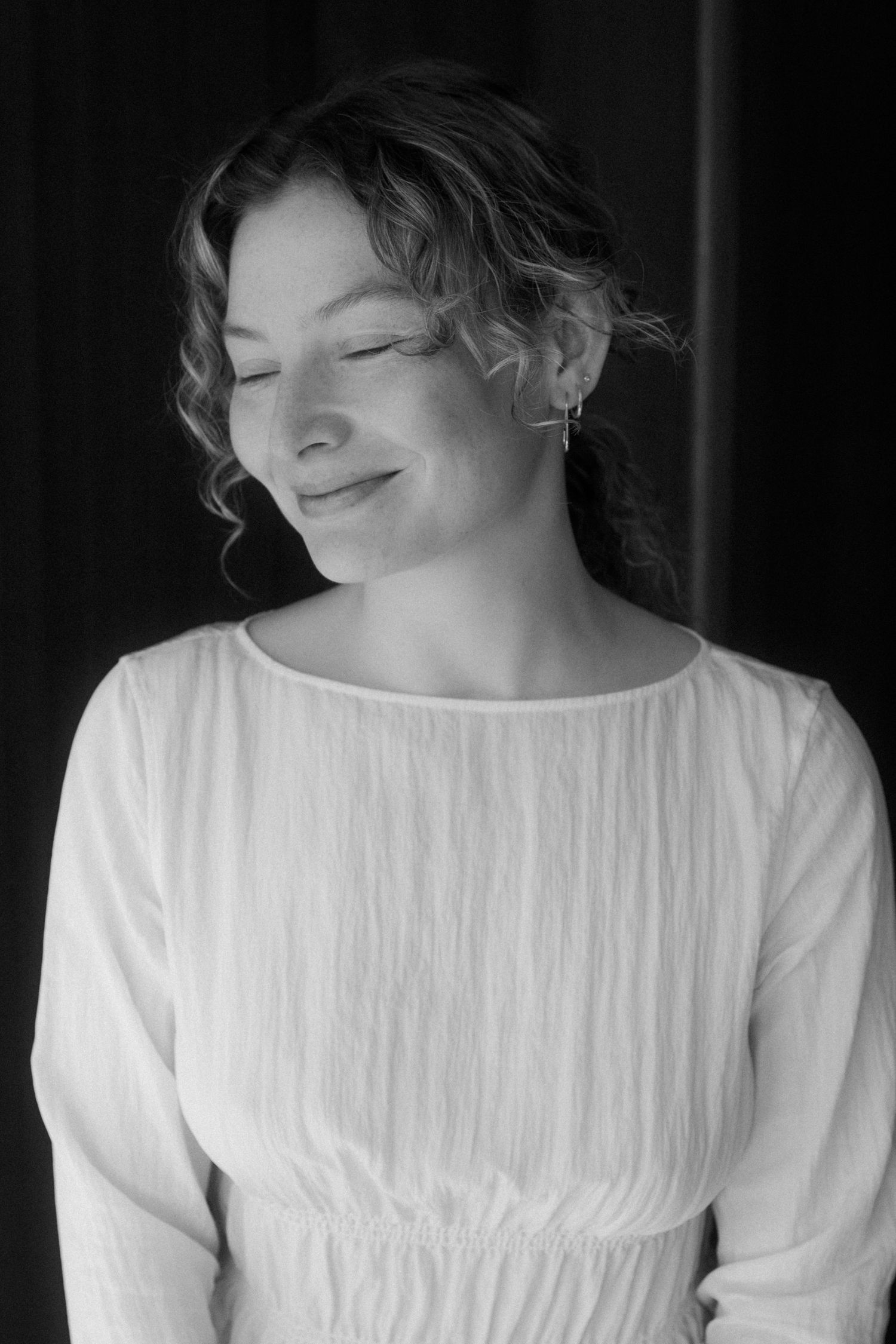 Black and white portrait of a person in a white dress with curly hair smiling with eyes closed.