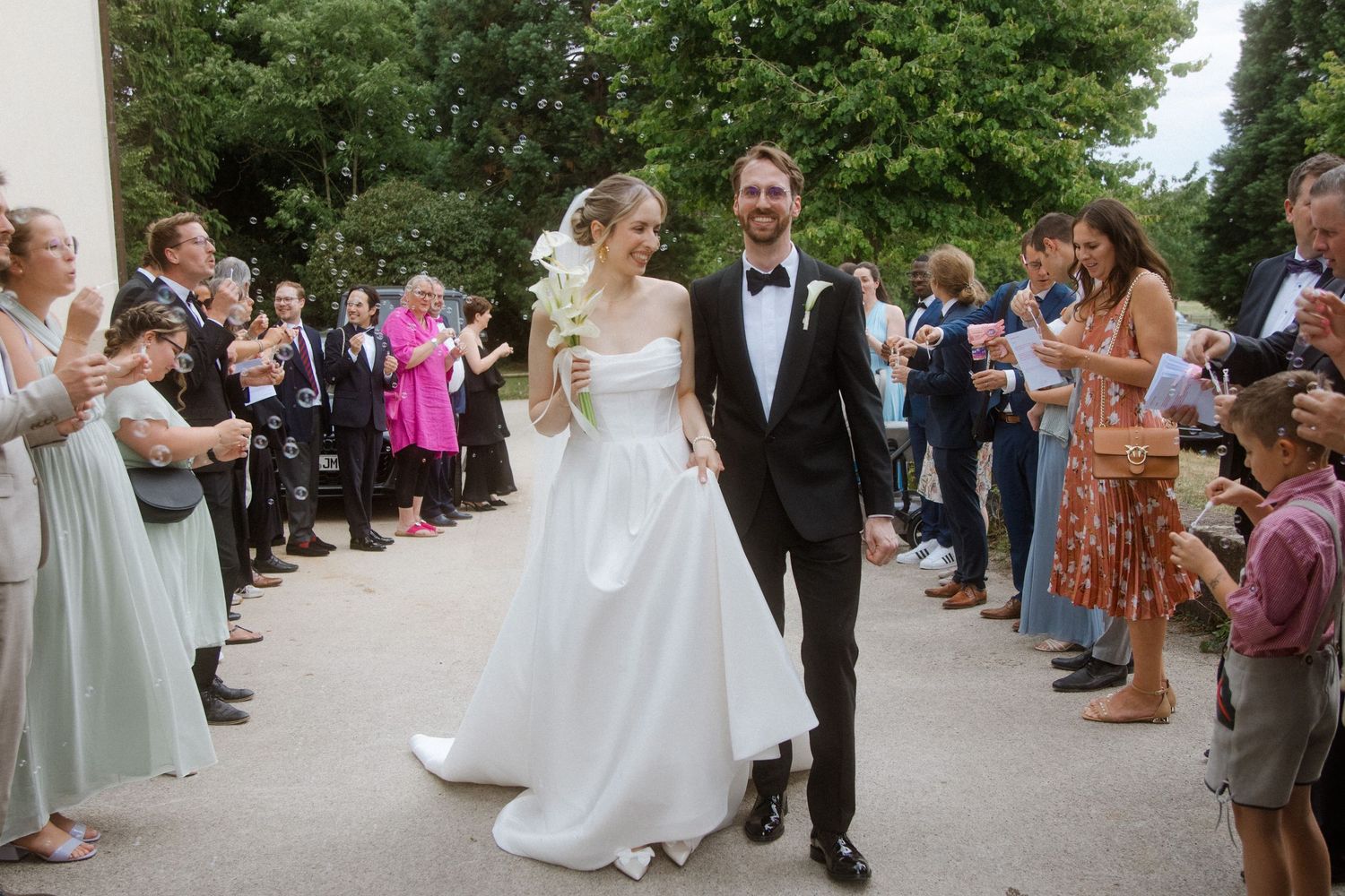 Wedding couple walks through crowd of guests on tree-lined outdoor path.