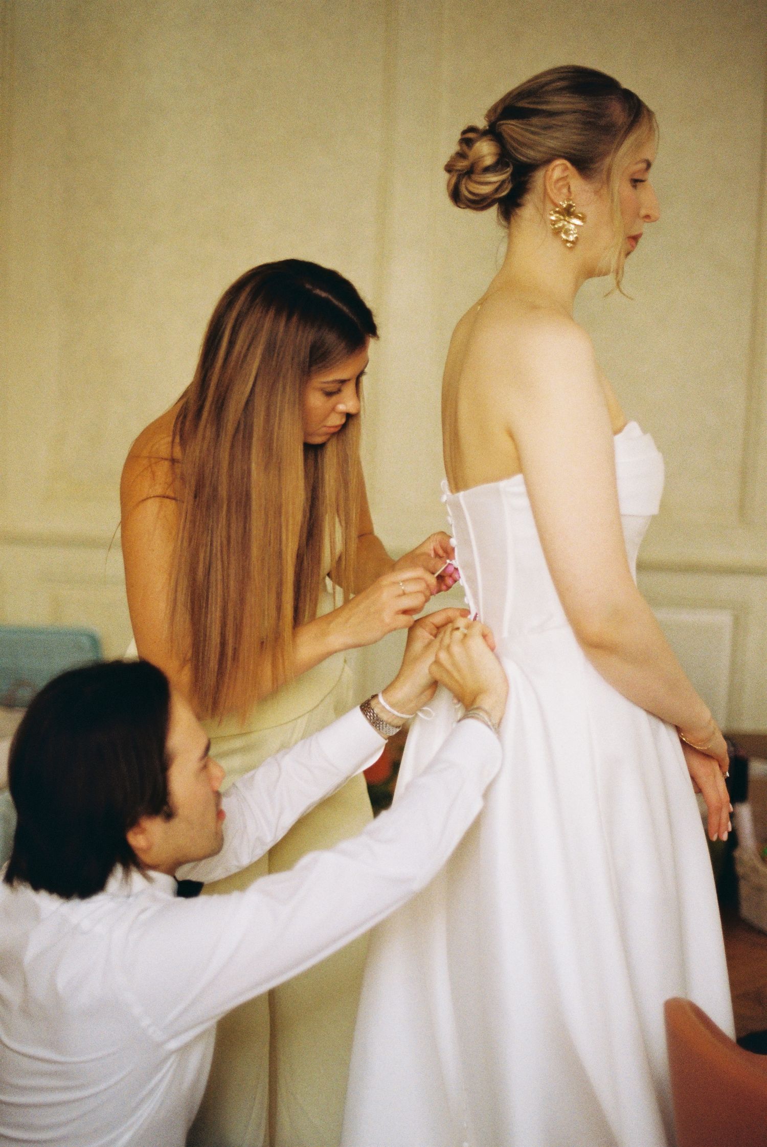 Two assistants helping to lace up a white wedding gown during final fitting.