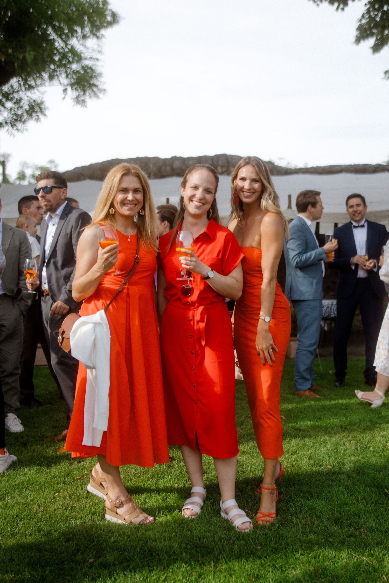 Three guests in vibrant coral outfits pose with drinks at an outdoor wedding reception.