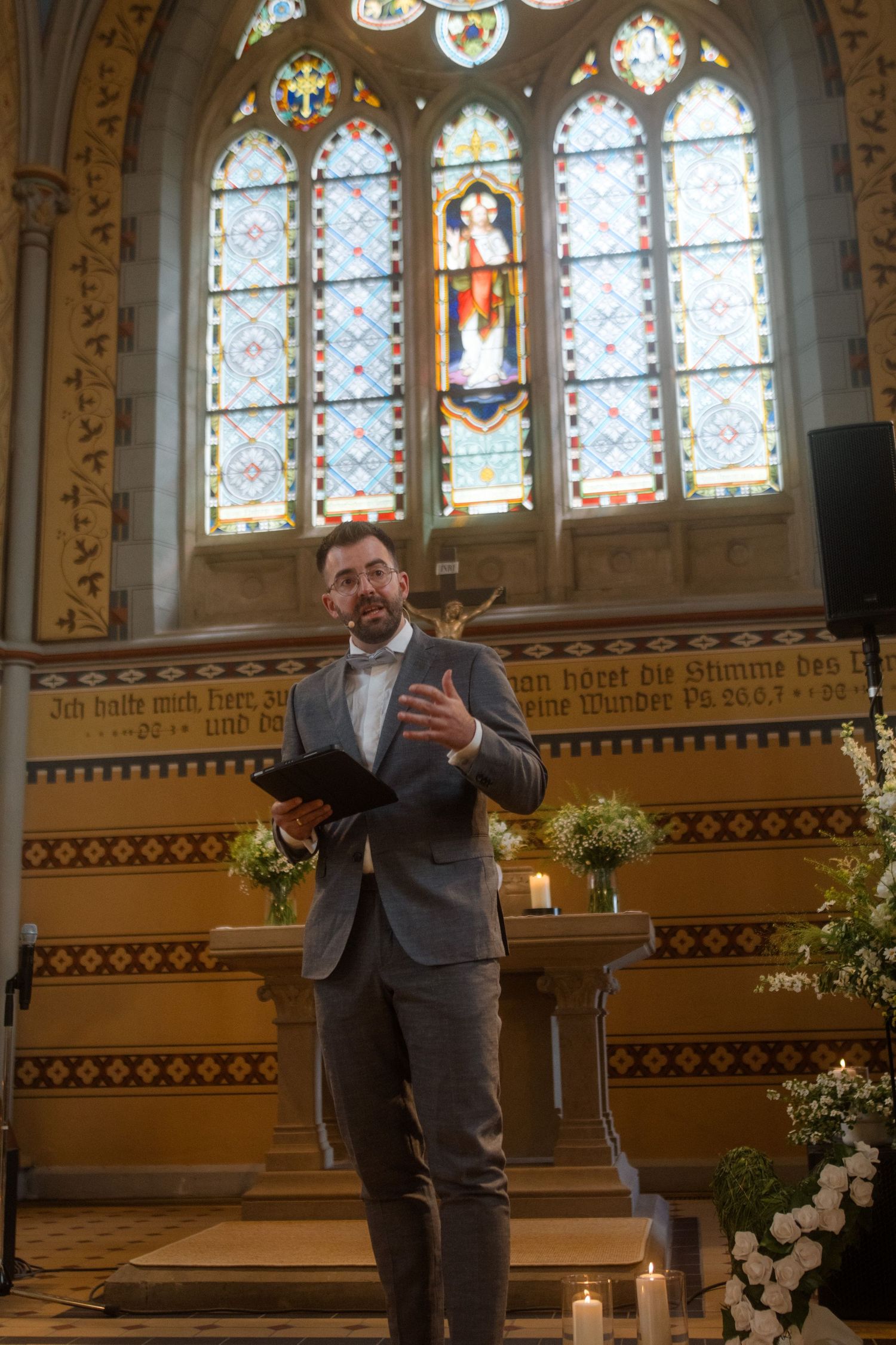 Speaker stands at church podium beneath colorful stained glass windows and decorative wall details.