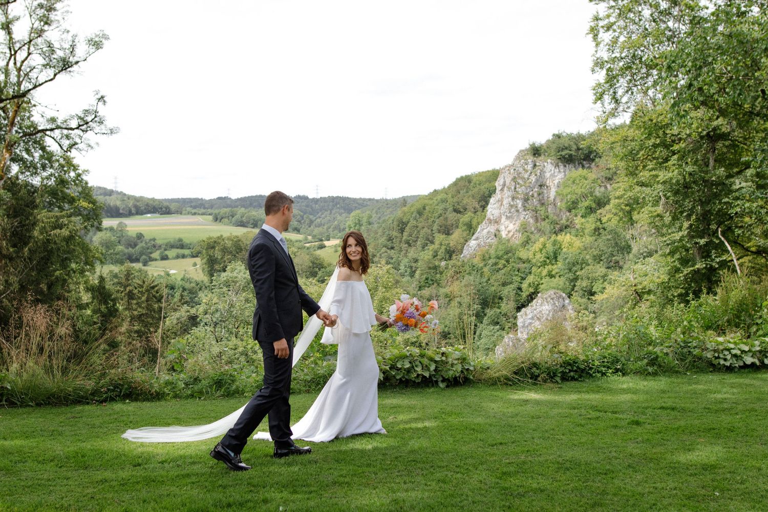 Wedding couple walks together on a hilltop lawn with dramatic cliff views in the background.