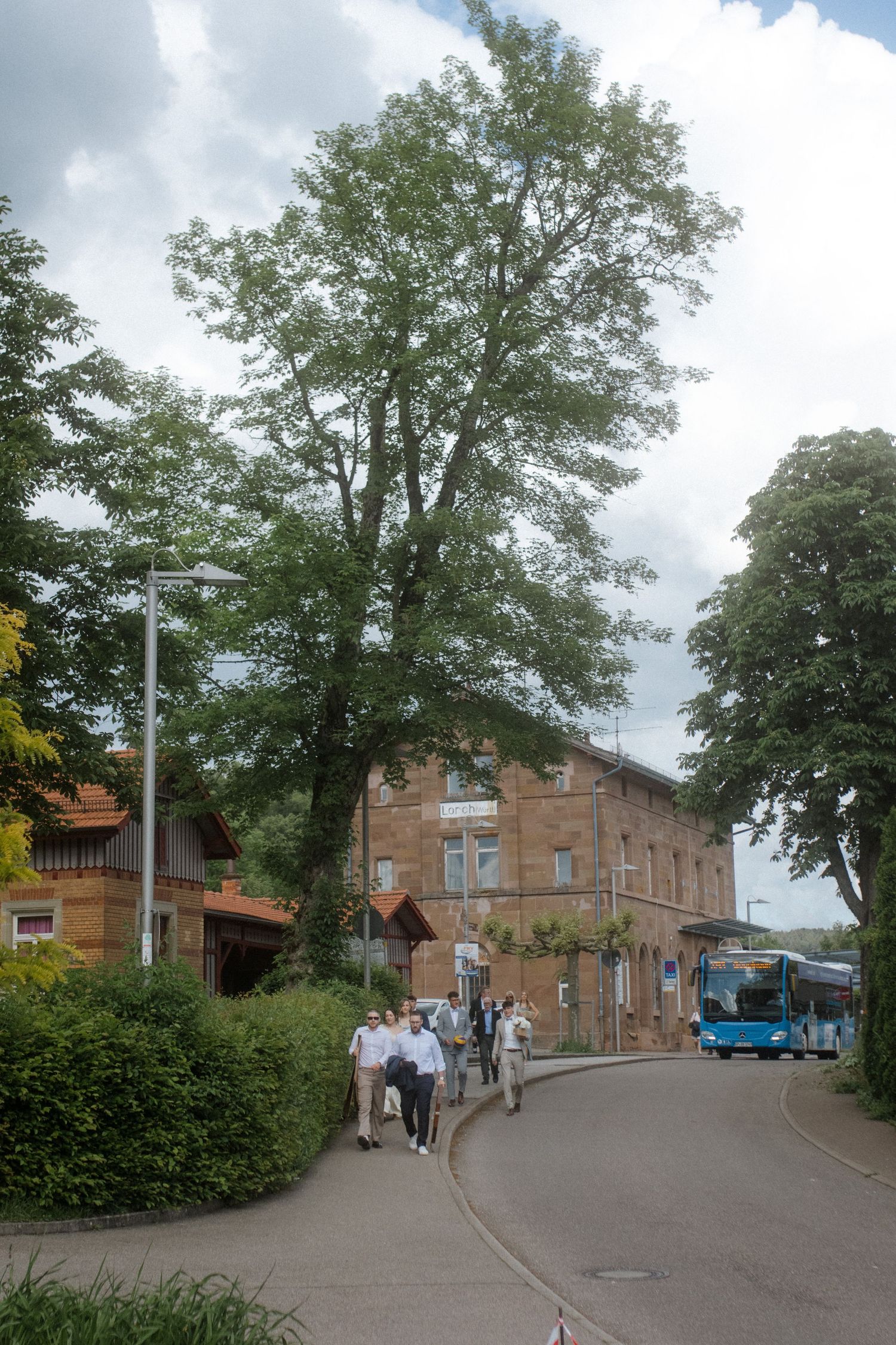Brick building on a curved street with trees and a blue bus in the background.