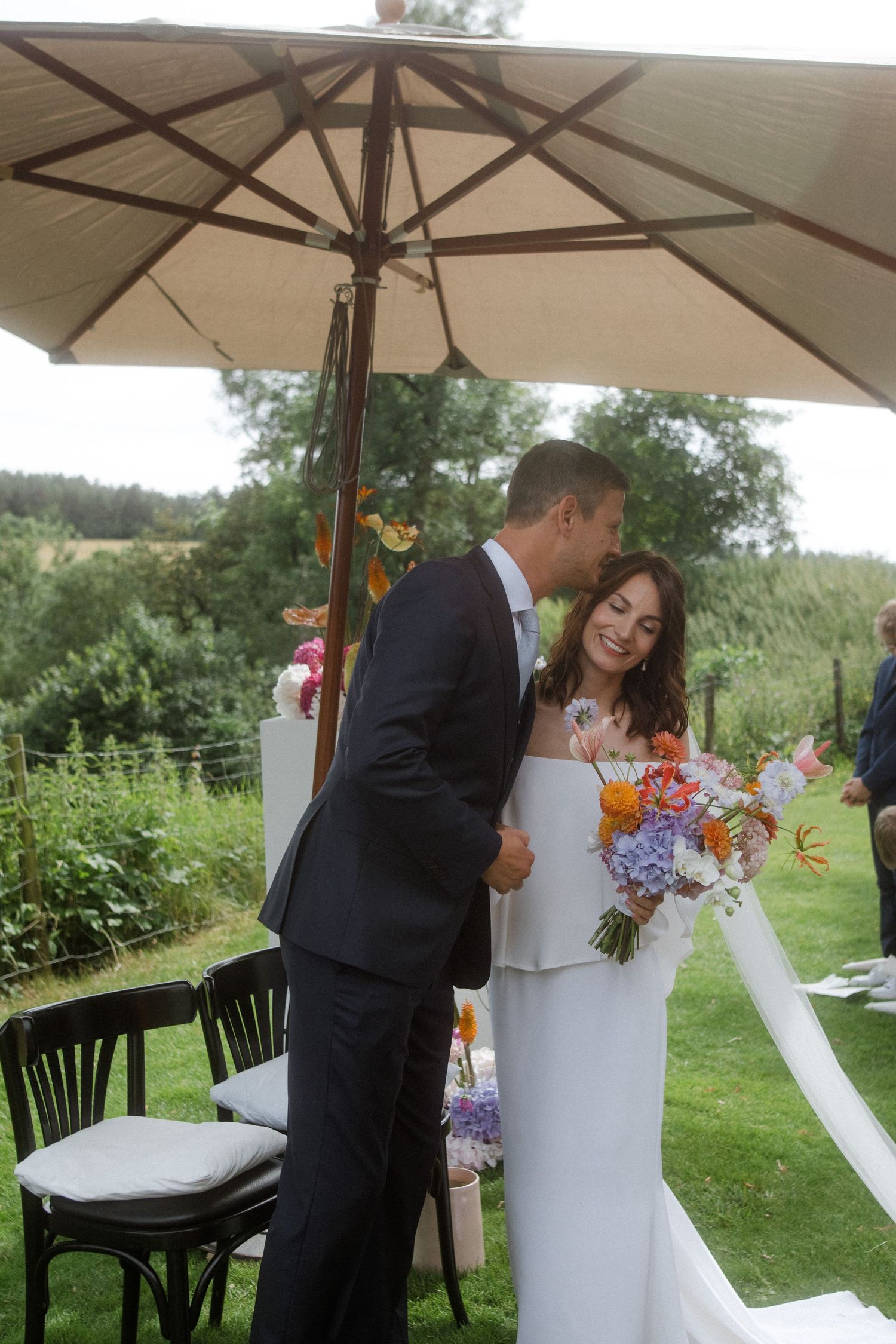 Wedding couple sharing intimate moment under beige umbrella in garden setting.