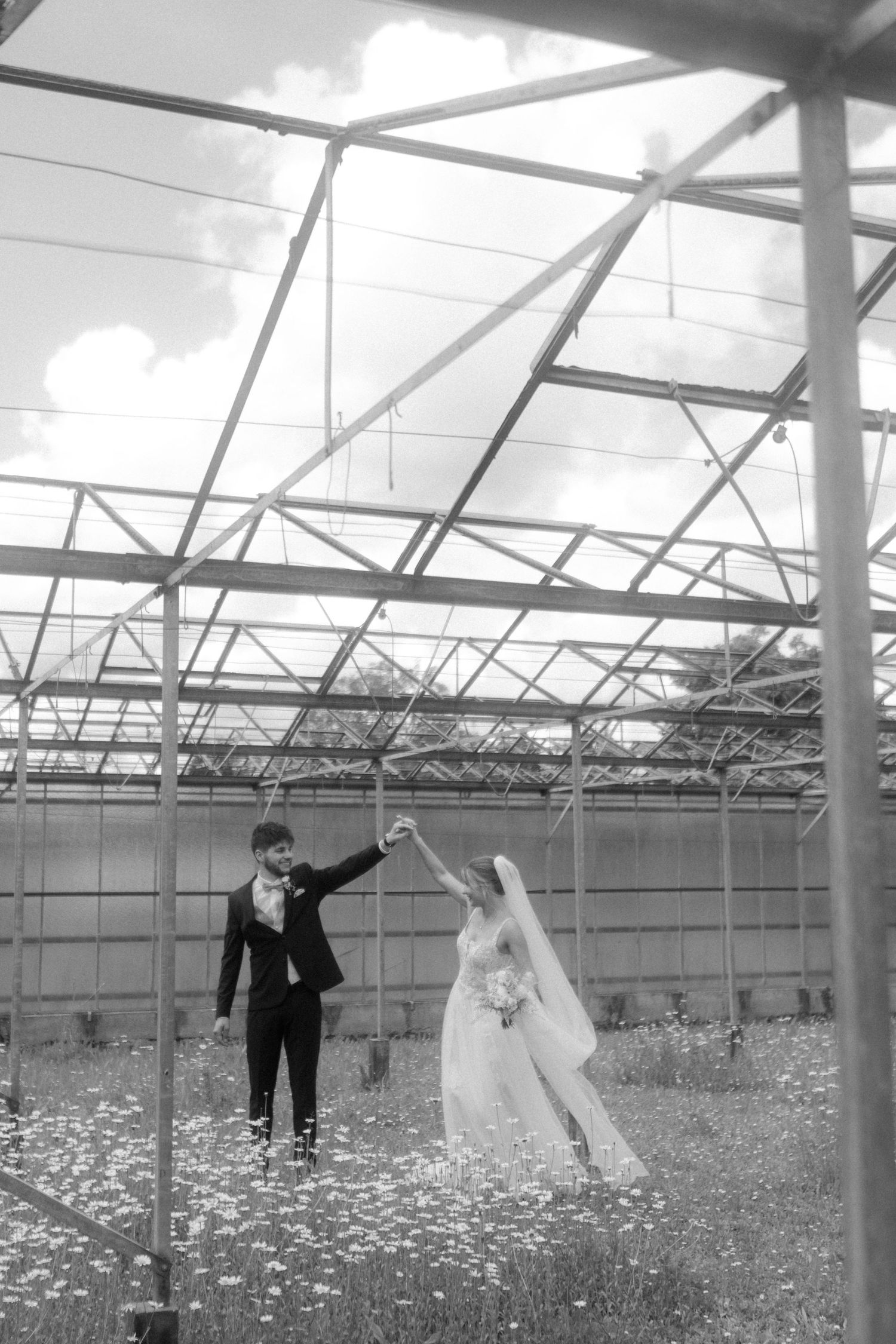 Black and white photo of wedding couple dancing playfully in a greenhouse setting.