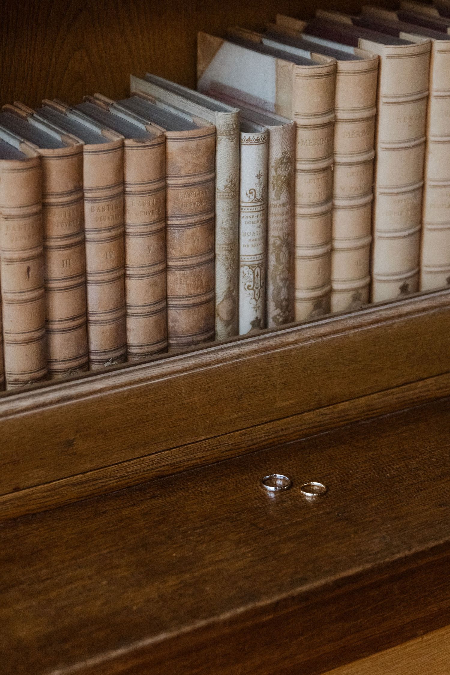Row of antique leather-bound books on wooden shelf with wedding rings below.