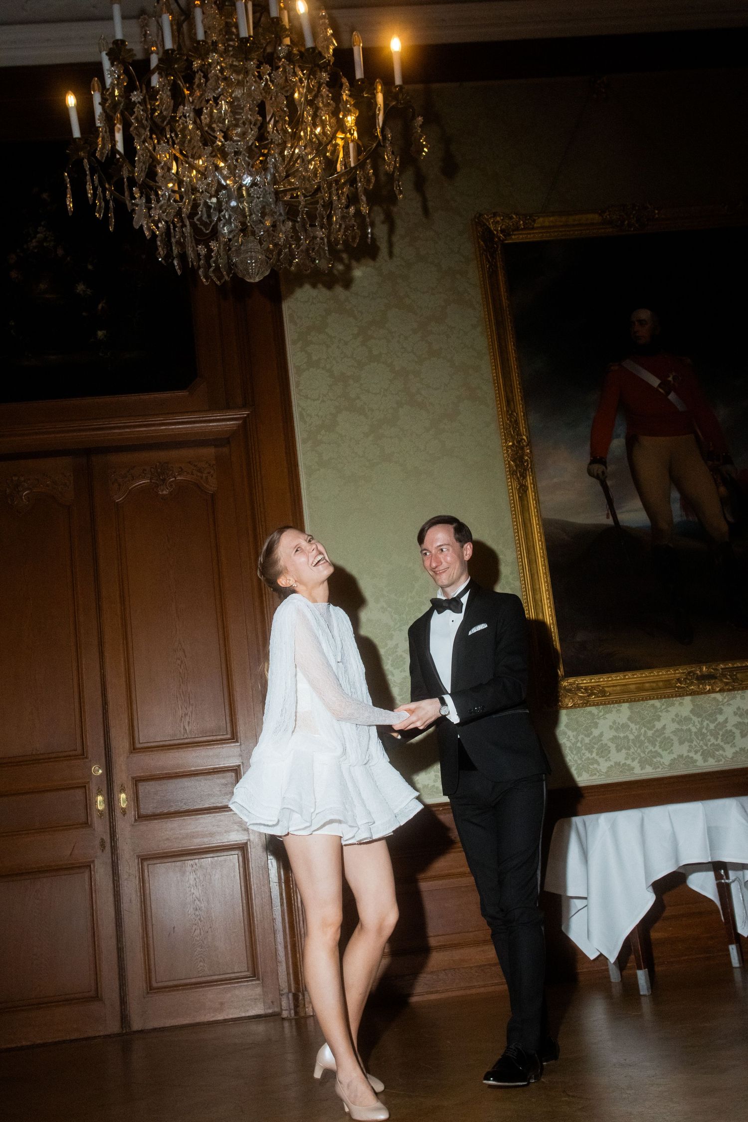 Couple dancing beneath a crystal chandelier in an ornate room with historic portraits.