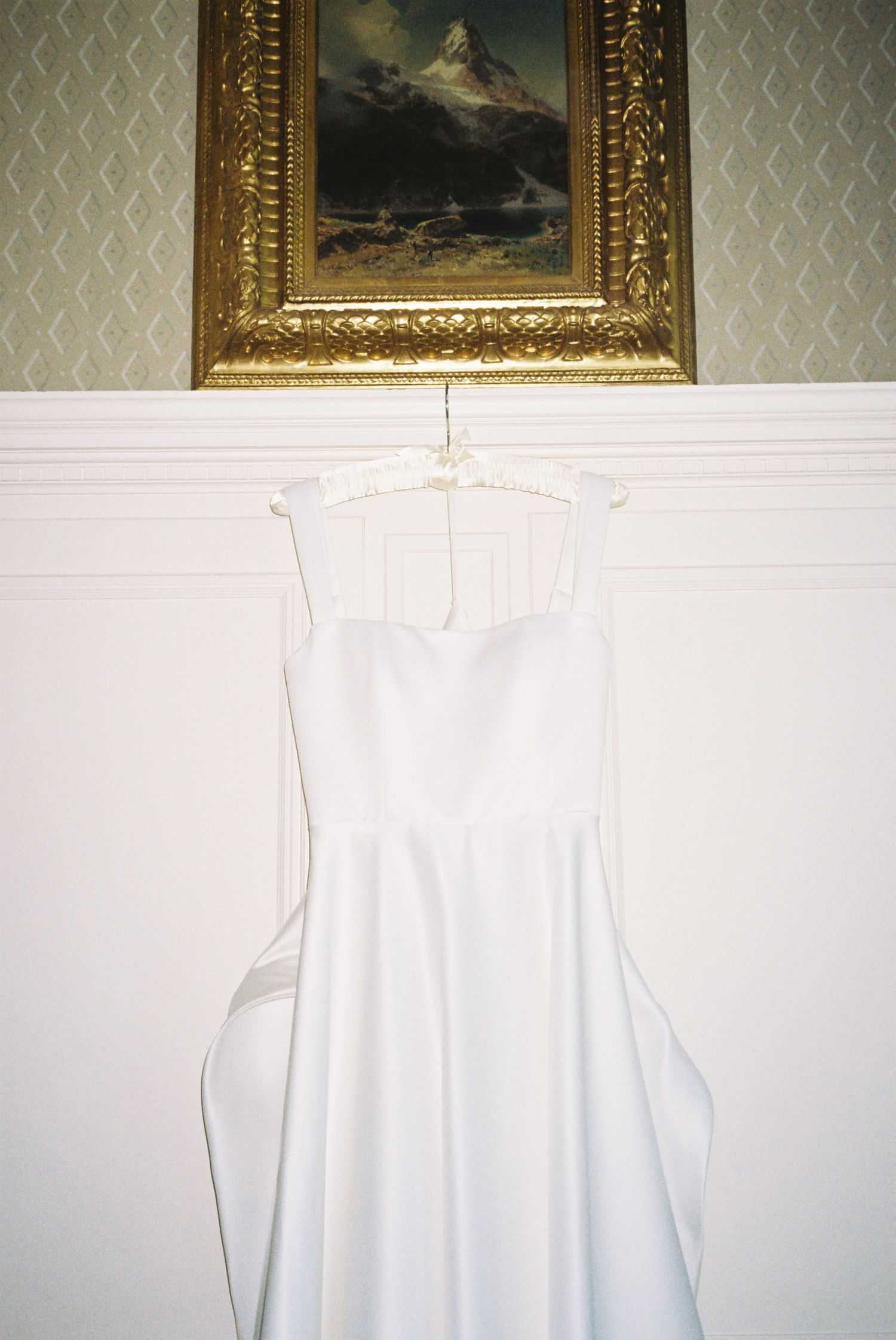 White wedding dress hanging against wall with ornate gold-framed paintings above.