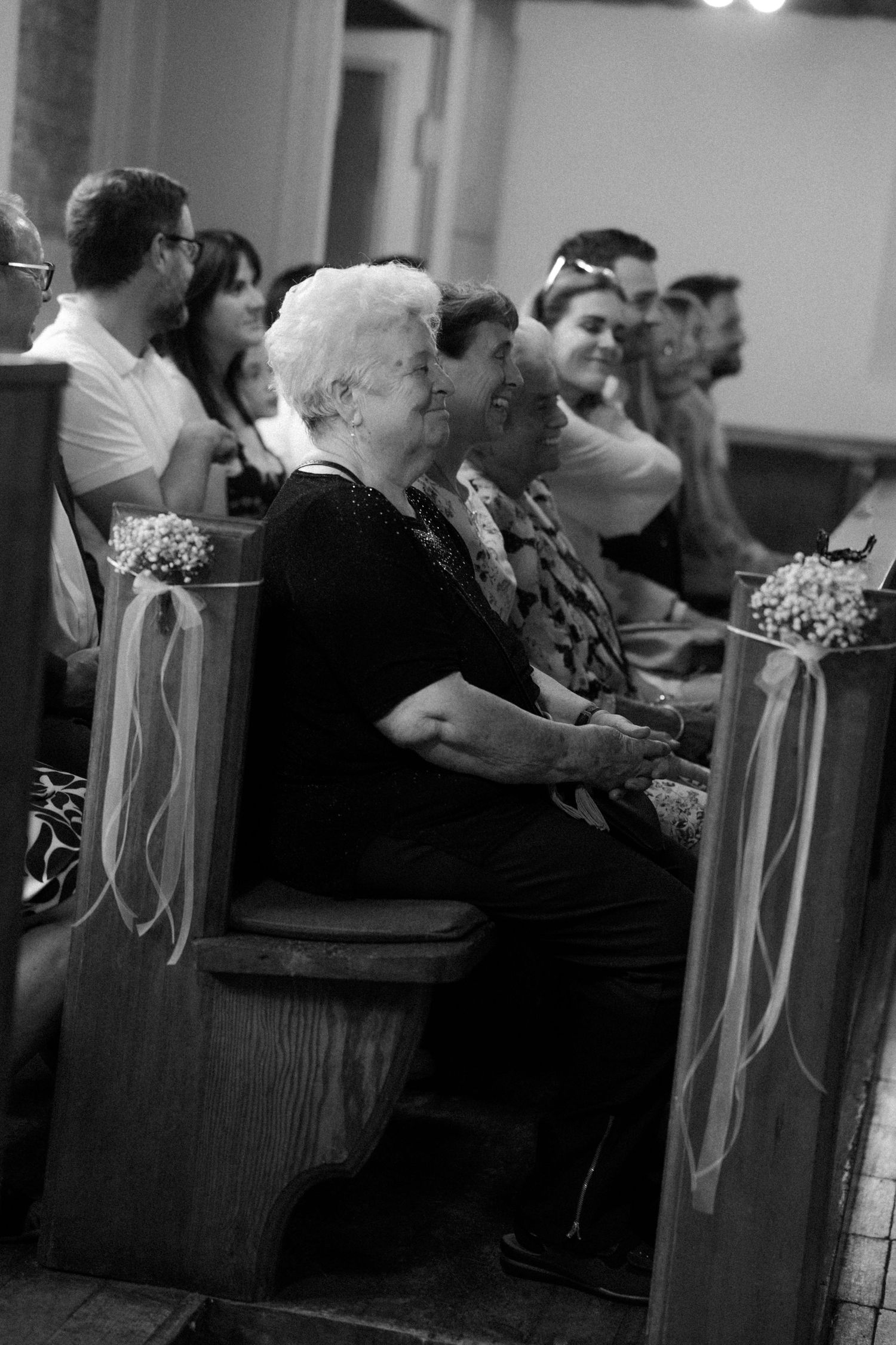 Wedding guests seated in church pews decorated with white ribbons and flowers during ceremony.