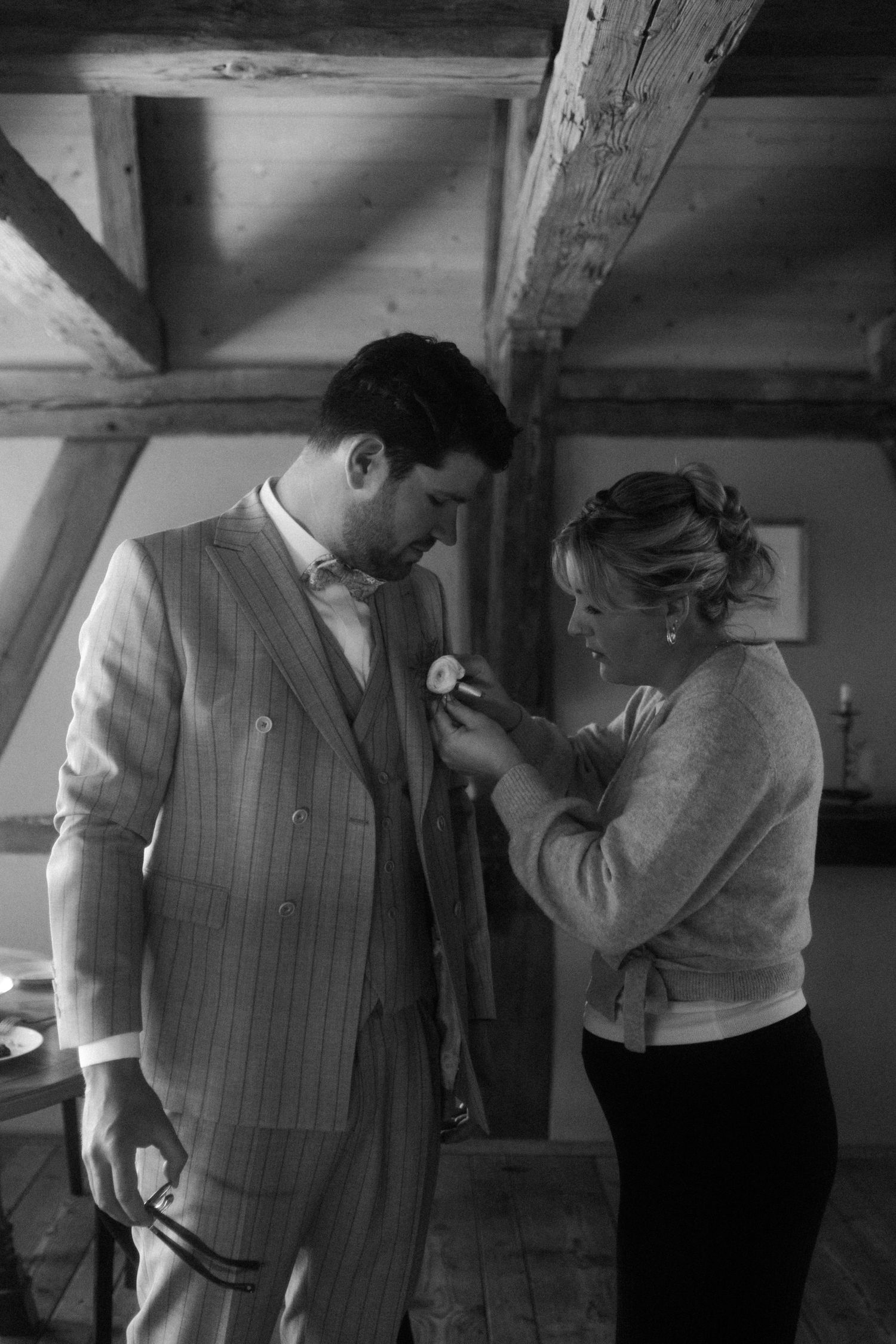 Black and white photo of a woman pinning a boutonniere on a mans suit jacket indoors.