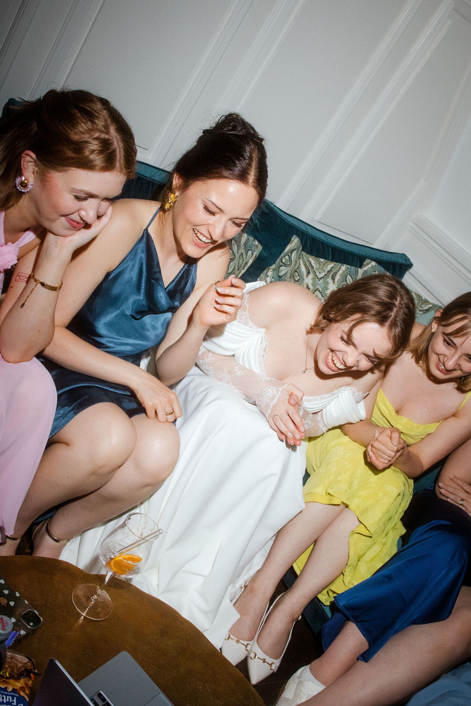 Bride in white dress laughing with bridesmaids in colorful dresses on a teal sofa.