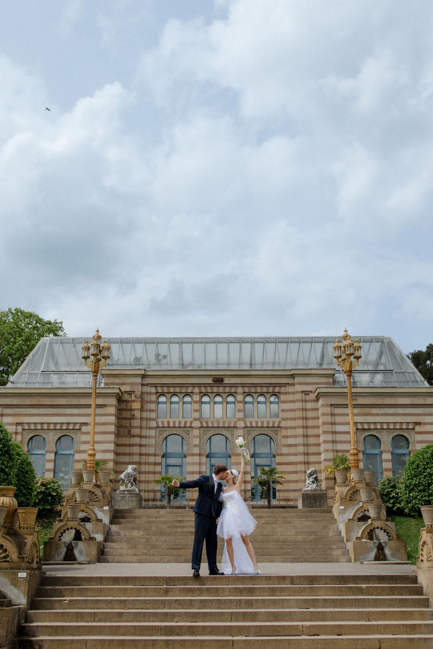 Newlyweds pose on grand stone steps leading to an elegant historic building with ornate architecture and decorative lamps.