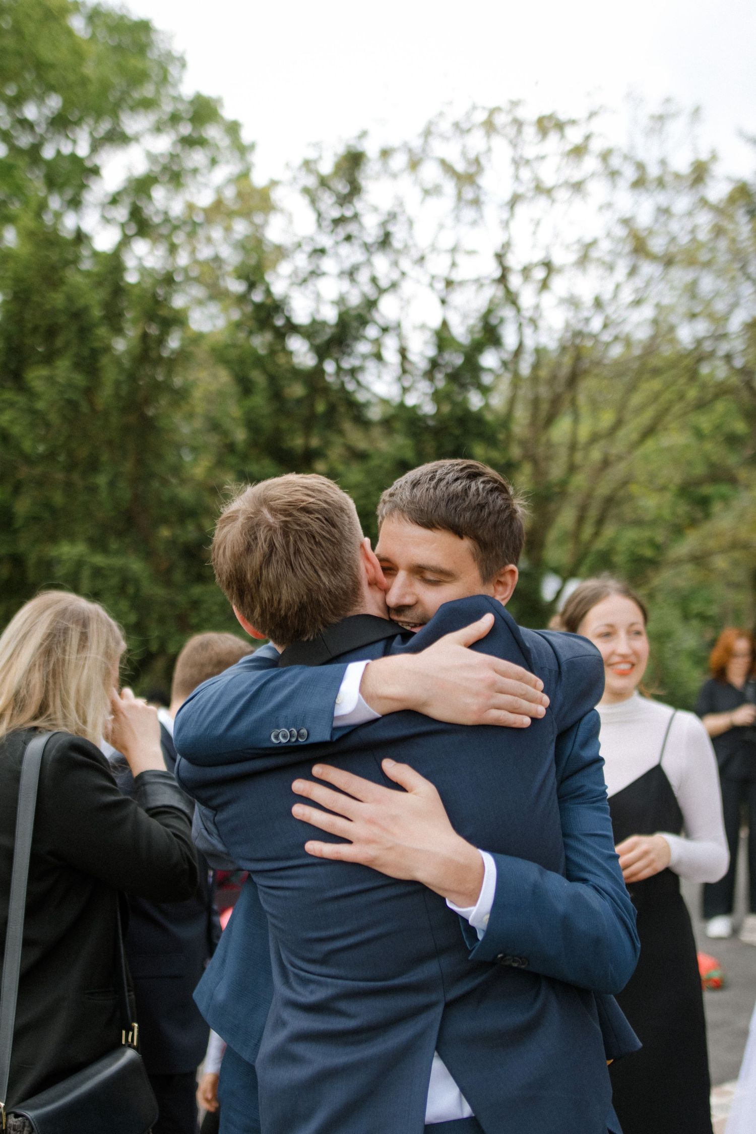 Two people in navy suits share an emotional embrace during an outdoor wedding celebration.