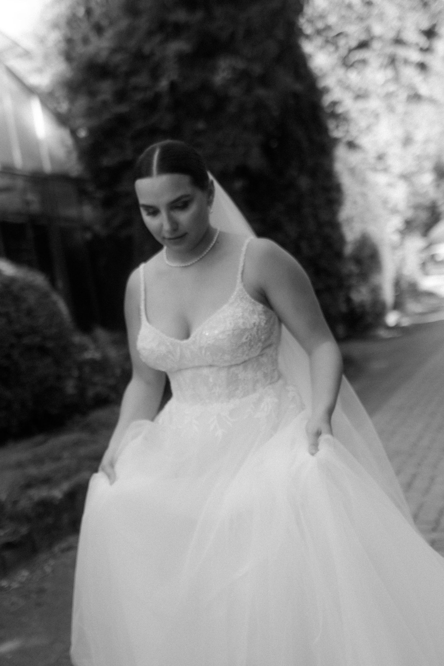 A black and white photo of a bride in a lace and tulle wedding gown looking down while adjusting her dress.