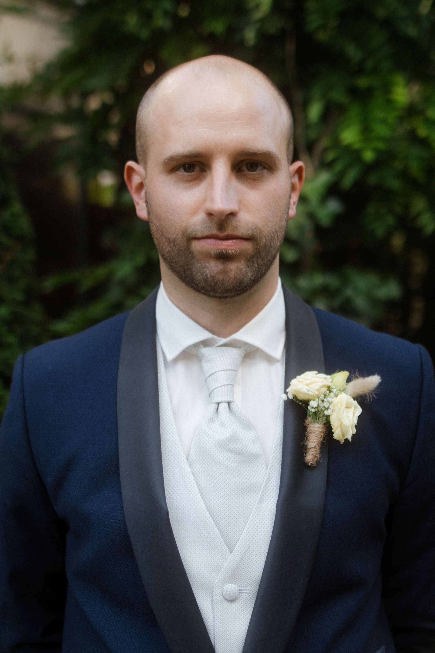 A formal portrait of a groom in a navy tuxedo with white vest and boutonniere.
