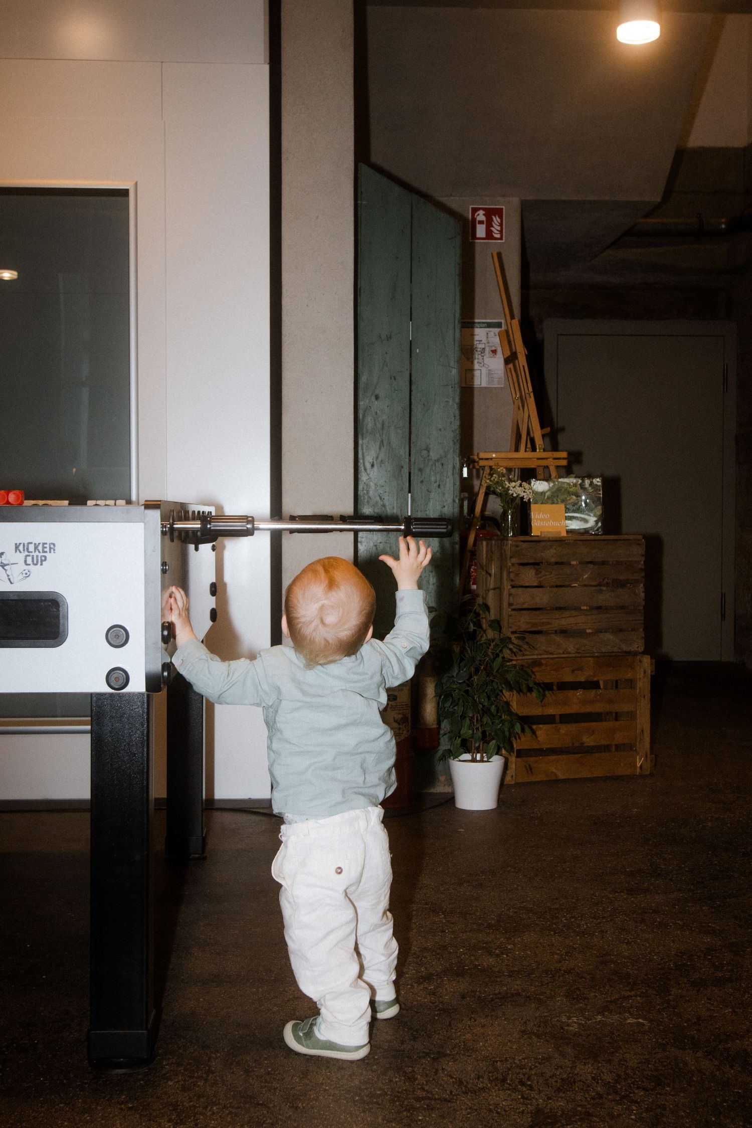 A small child in a gray sweater reaching up toward a vending machine in a dimly lit room with wooden crates.