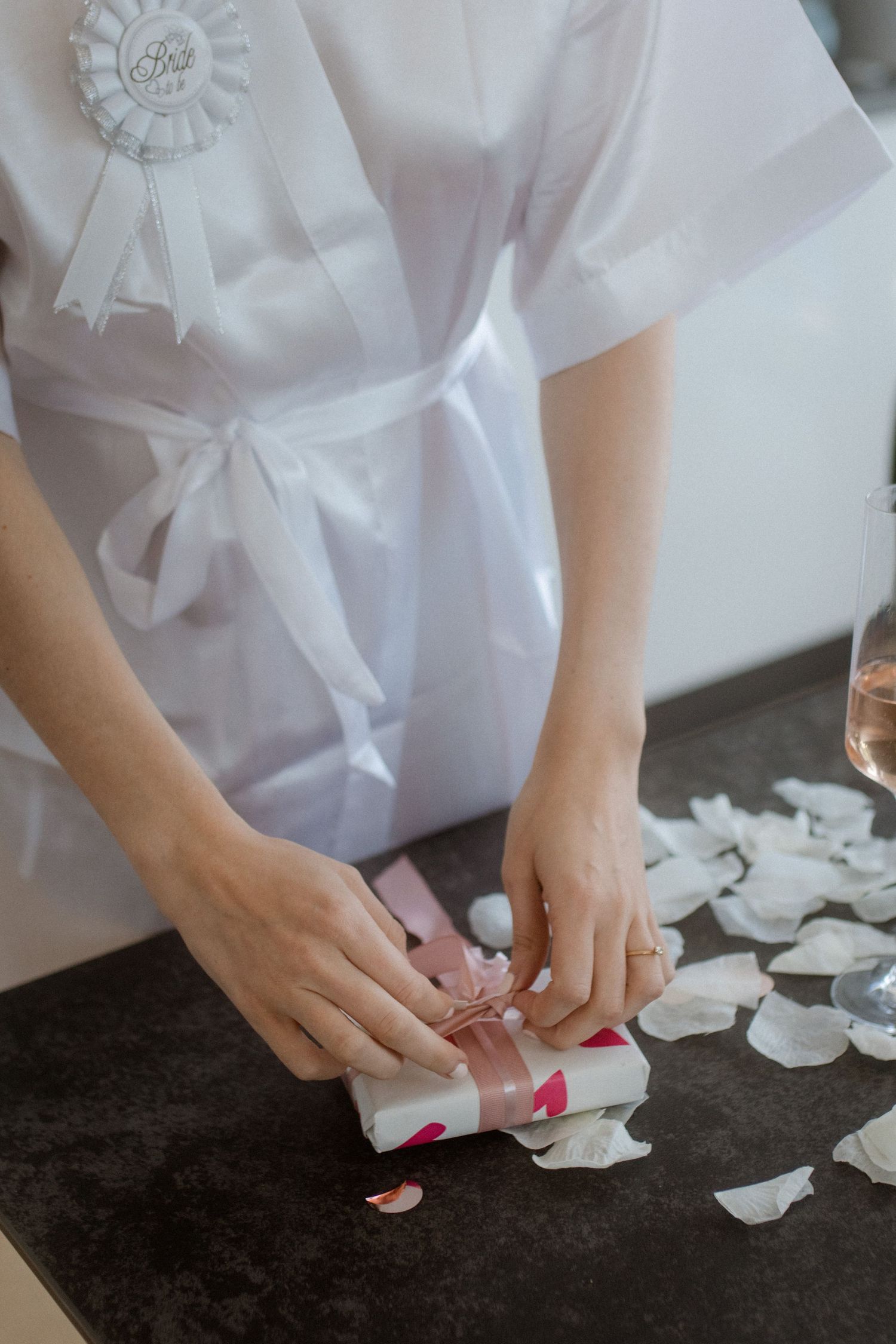 Close-up of hands with pink nails wrapping a small gift, wearing white satin robe.