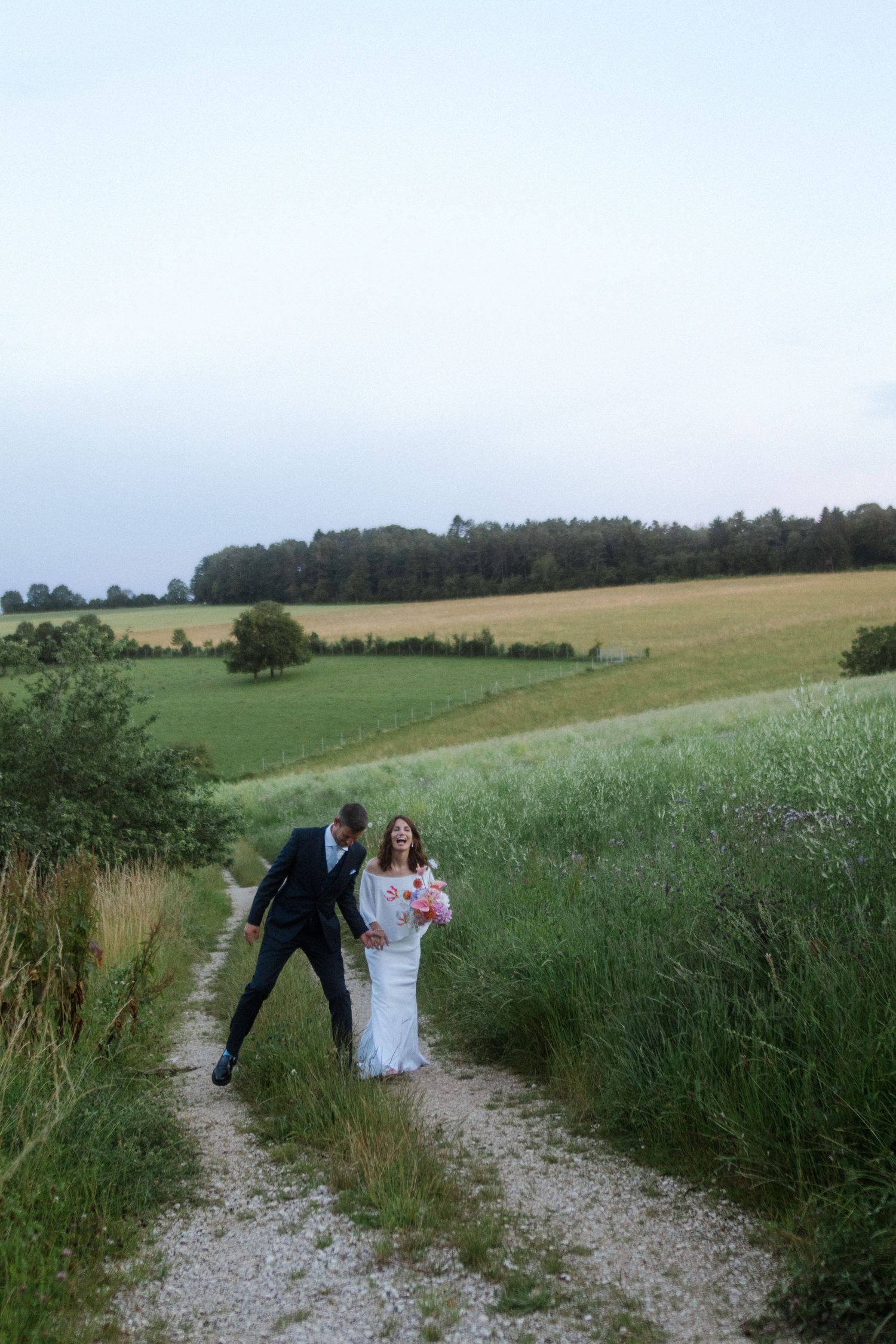Newlyweds walking together on a gravel path surrounded by lush summer fields.