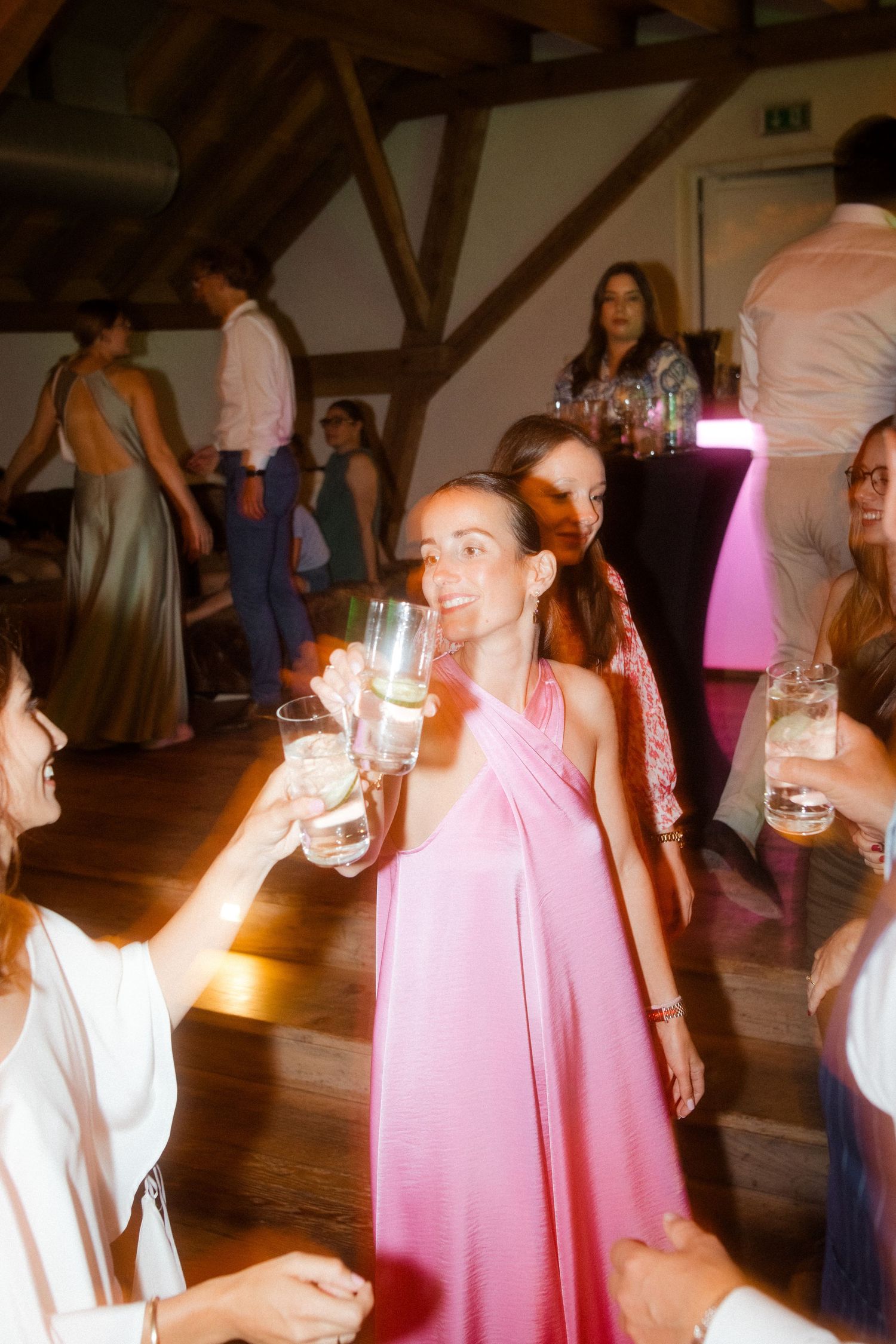 Wedding guests in pink dresses toasting with drinks on a wooden dance floor.
