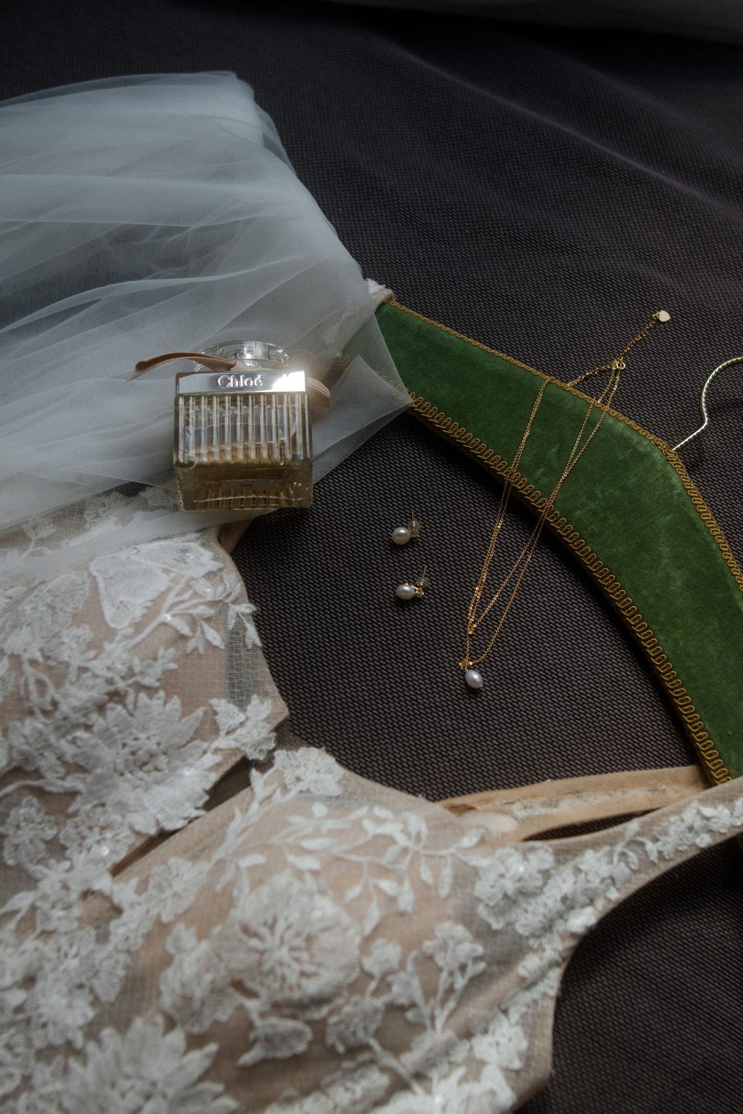 Wedding accessories including perfume, lace details, jewelry and veil arranged on dark fabric.