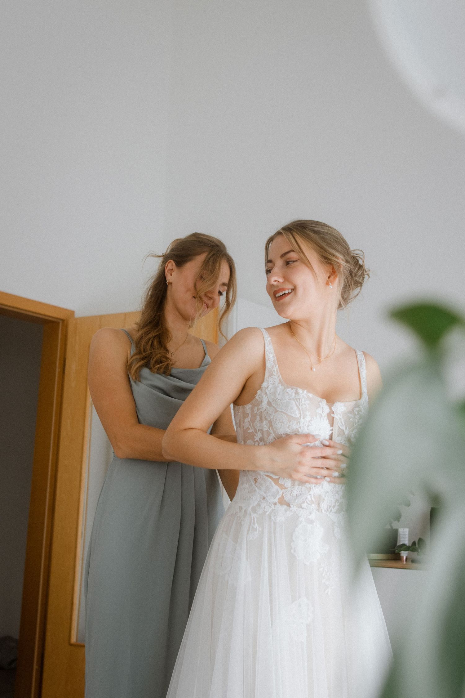 Two people share a joyful moment while preparing in a sunlit room, one wearing a white wedding gown.