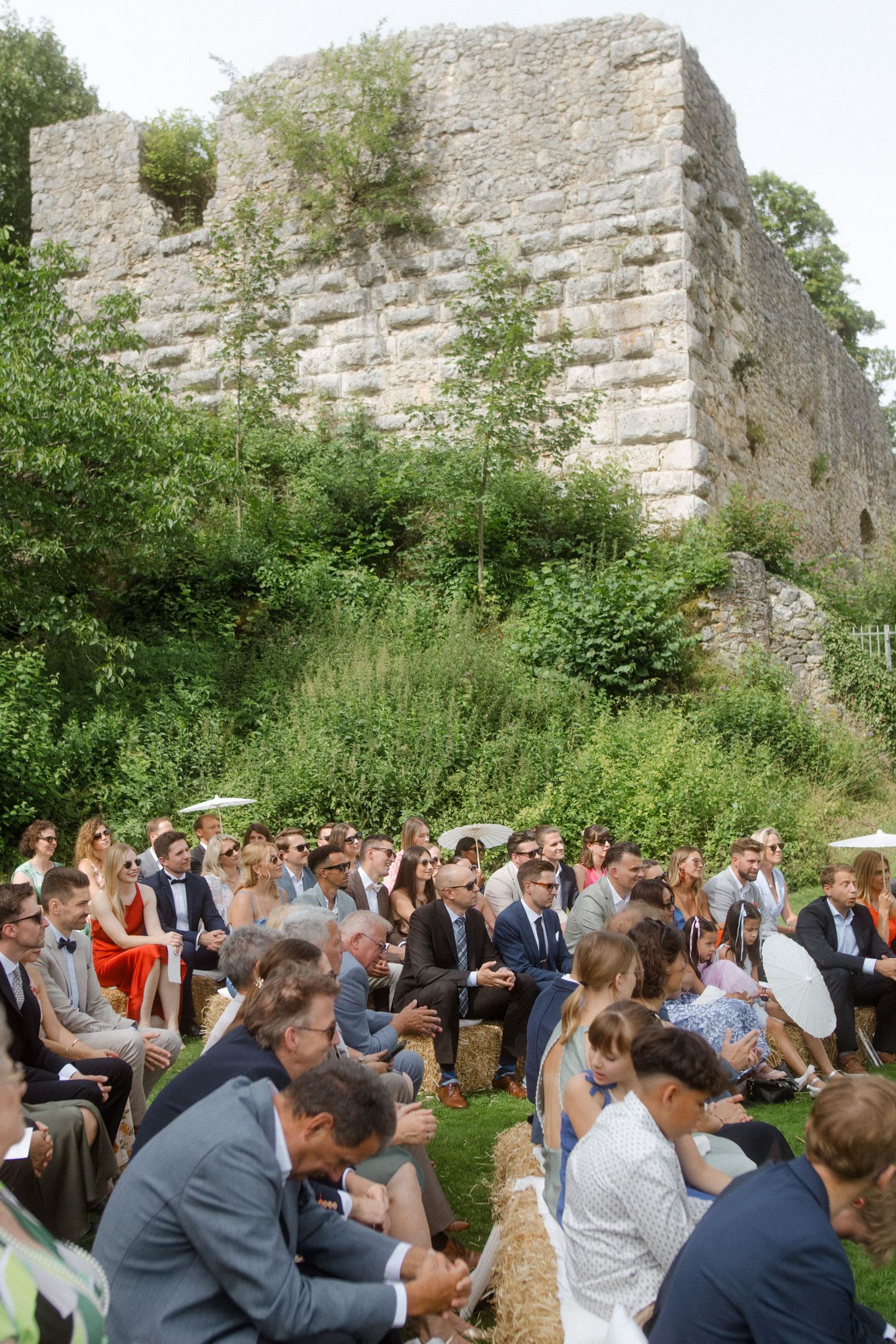Wedding guests seated on hay bales attend an outdoor ceremony in front of ancient stone castle walls.