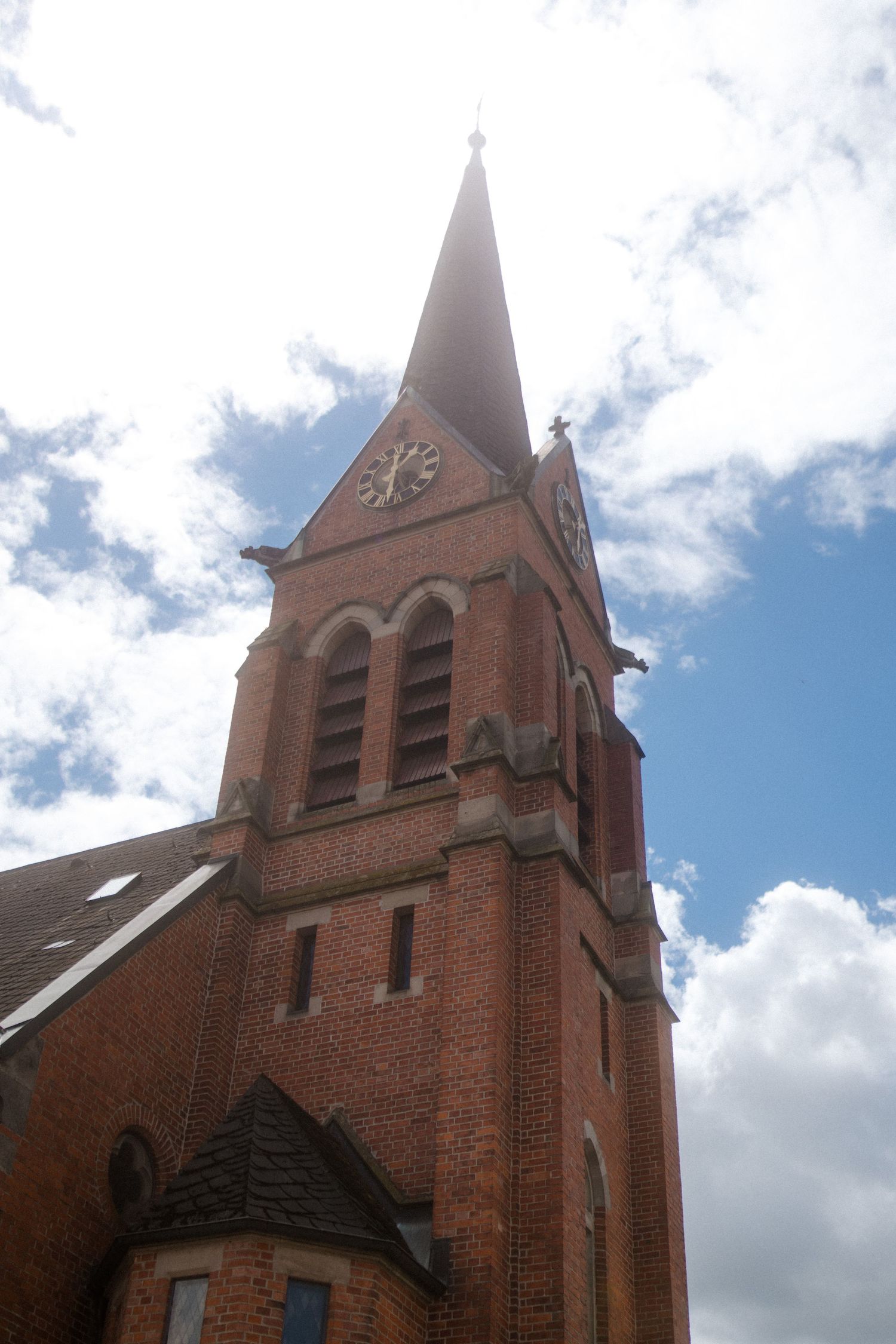 Tall brick church steeple with clock face rises against blue sky with white clouds.