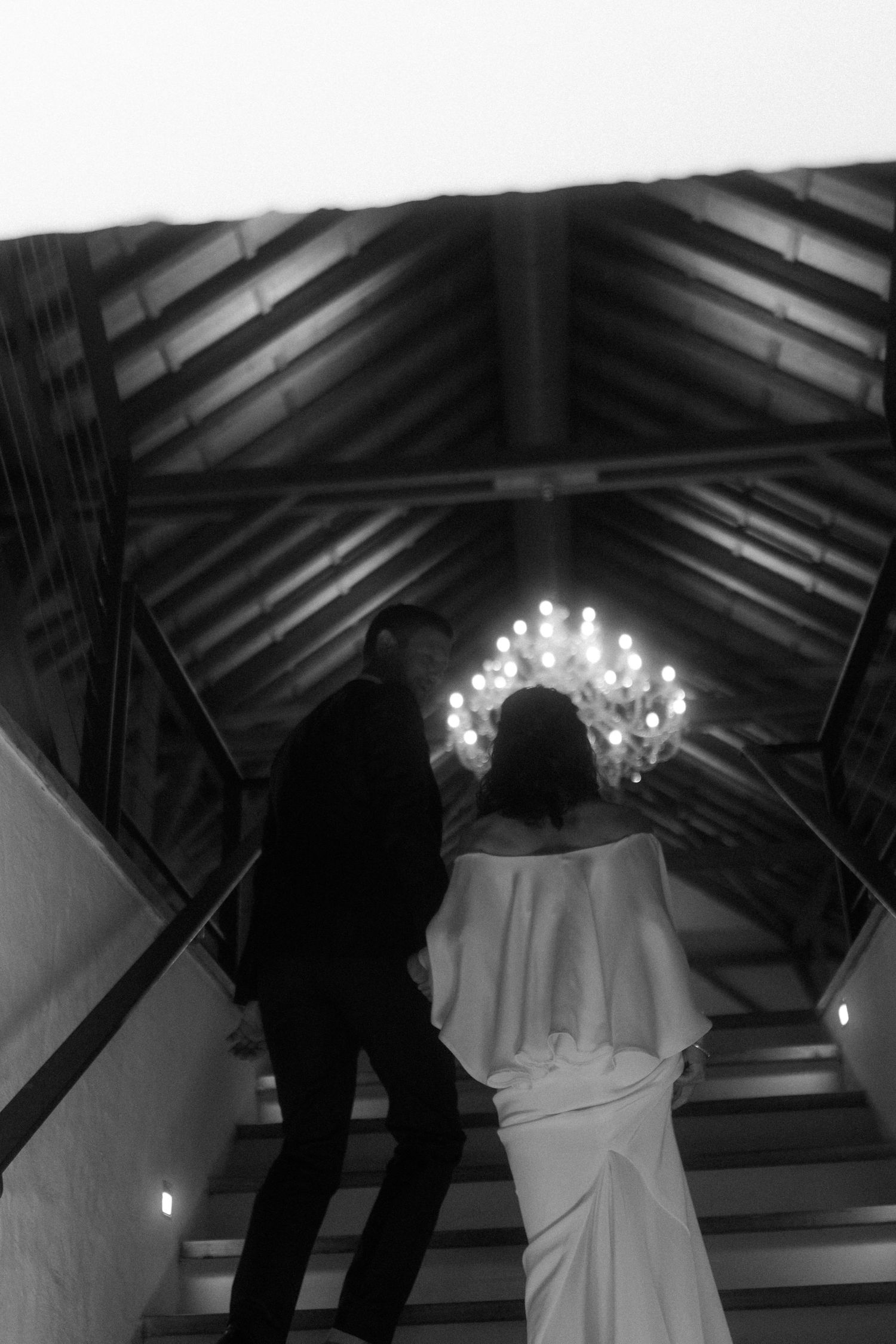 Black and white photo of couple ascending stairs under dramatic chandelier lighting.