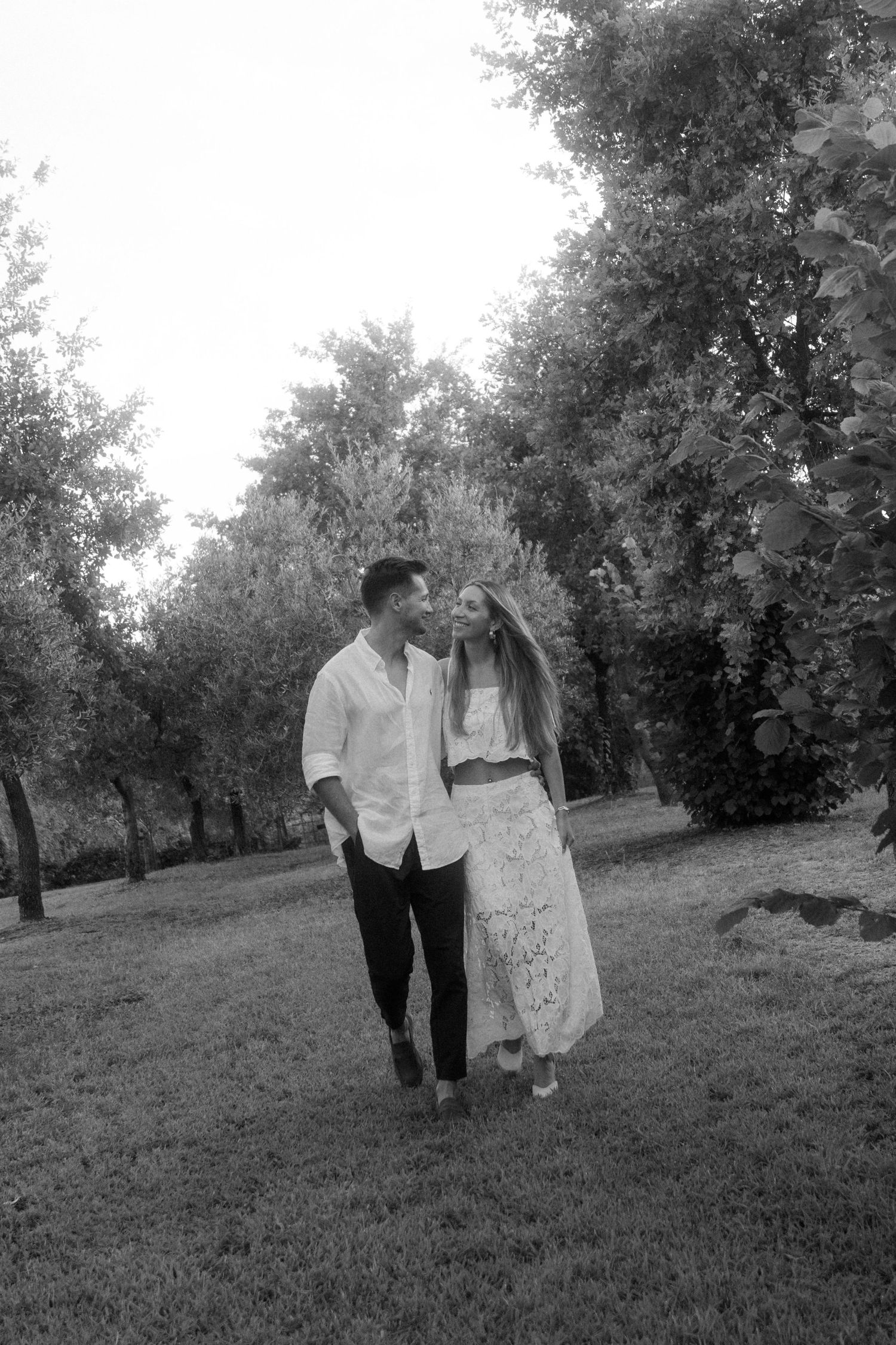 A romantic couple walks together through an orchard in black and white.
